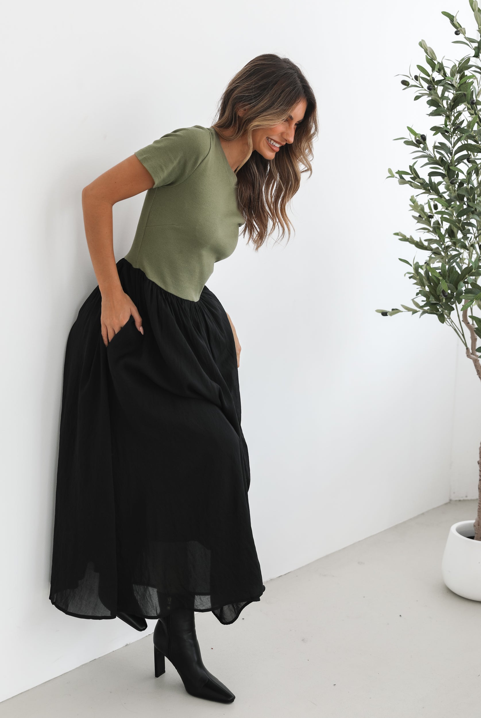 woman wearing a short sleeve round neck dress with green top and black flowy skirt paired with black boots in a white background studio