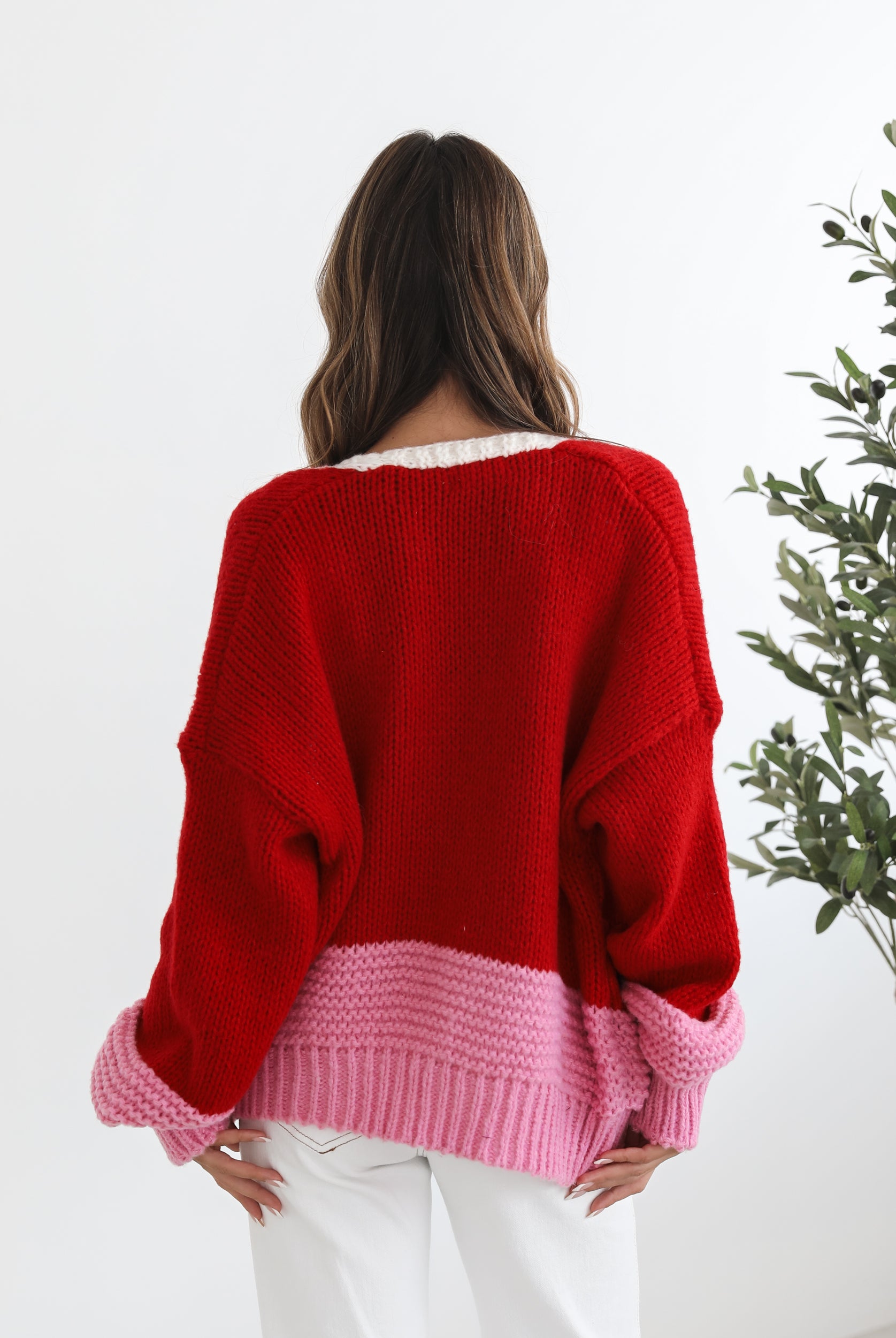 A woman sitting on a chair wearing a red, pink, and white striped cardigan with long sleeves and cuffs, paired with a white top and white pants.