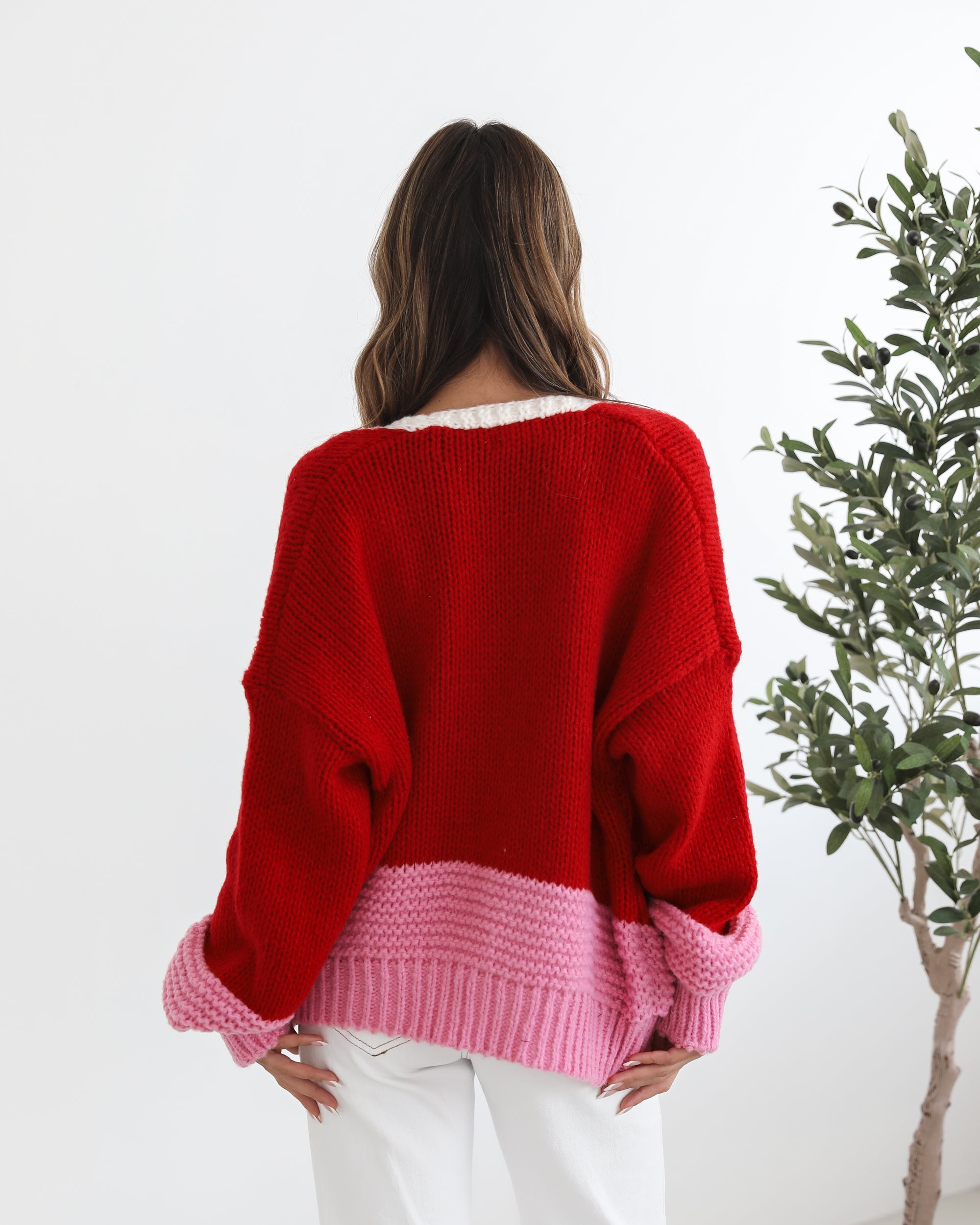 A woman sitting on a chair wearing a red, pink, and white striped cardigan with long sleeves and cuffs, paired with a white top and white pants.
