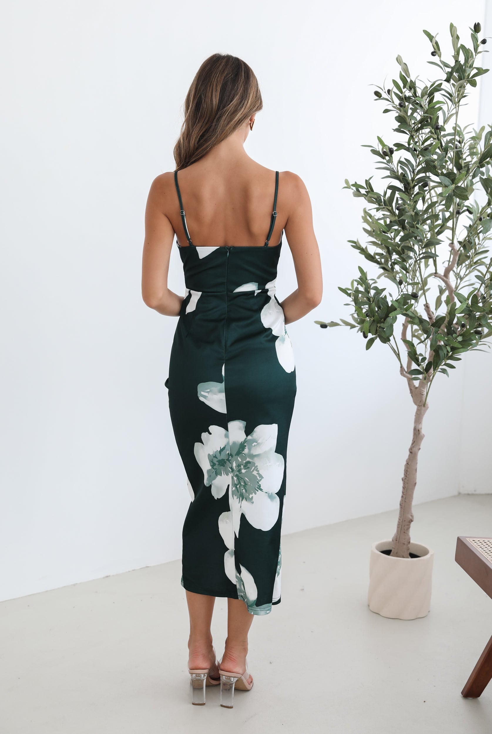 A woman wearing an emerald floral dress with spaghetti straps, a v-neckline, and an overlap skirt with a high-low hemline. She is standing next to a potted plant.