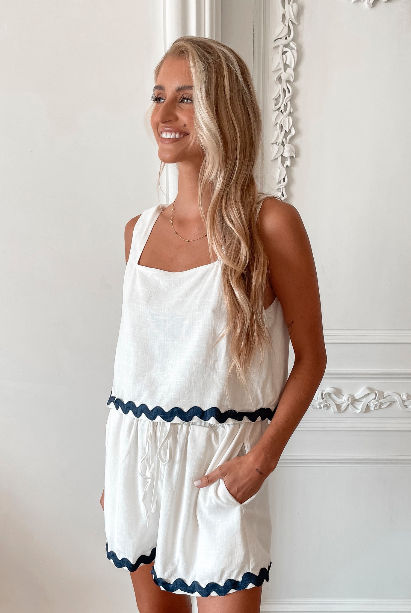 A woman is standing, wearing white high-waisted shorts with a string detail and side pockets, paired with a sleeveless top.