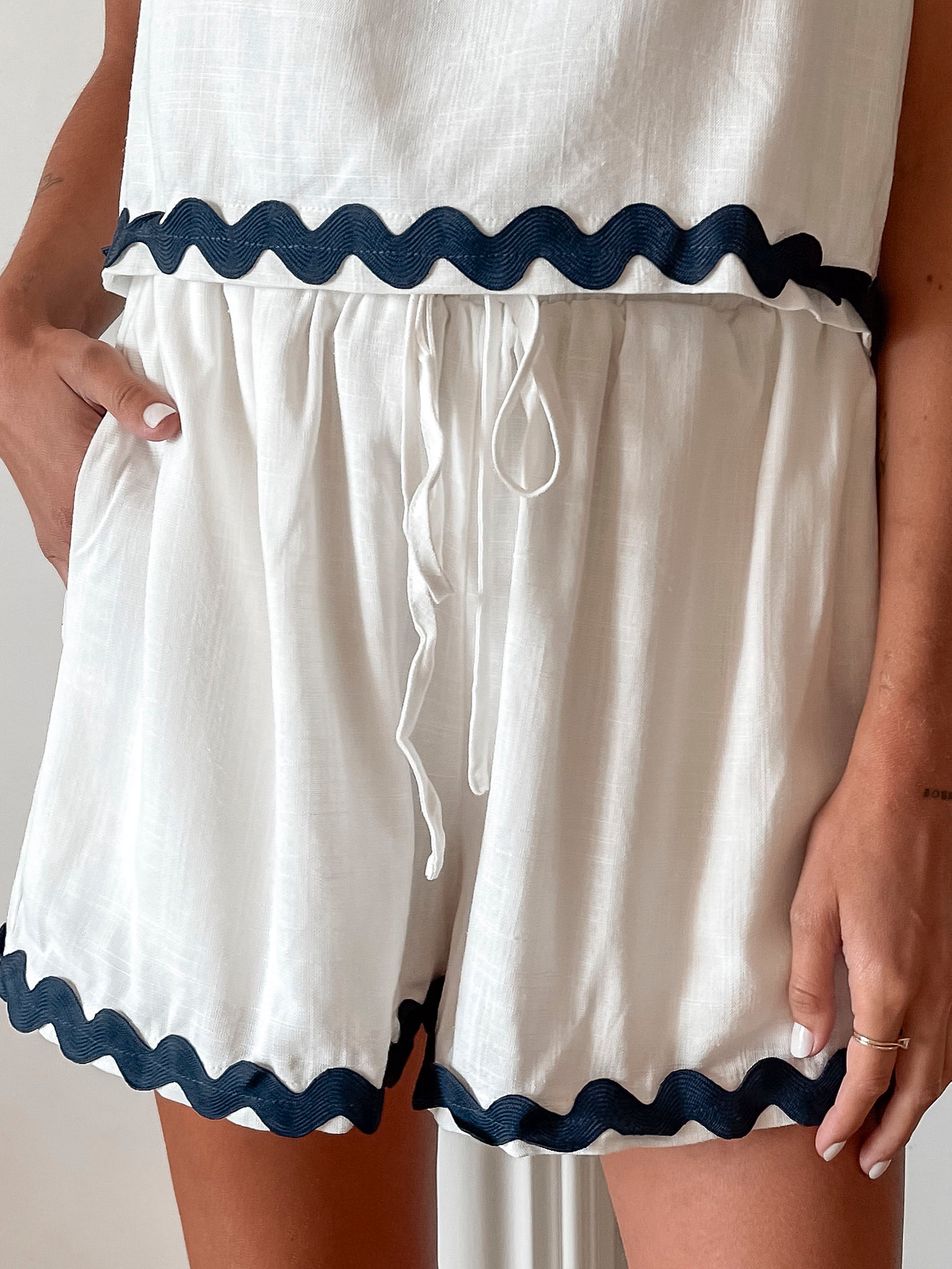 A woman is standing, wearing white high-waisted shorts with a string detail and side pockets, paired with a sleeveless top.