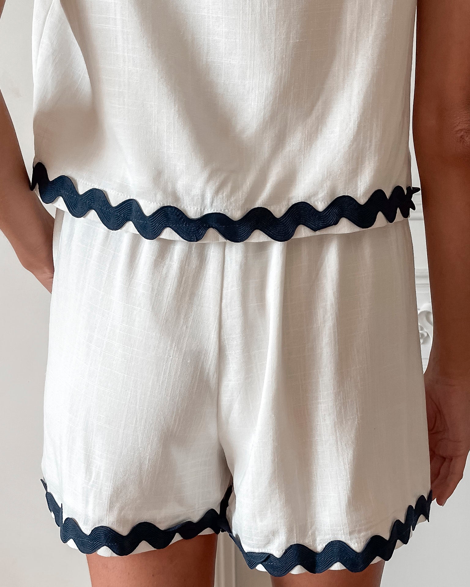 A woman is standing, wearing white high-waisted shorts with a string detail and side pockets, paired with a sleeveless top.