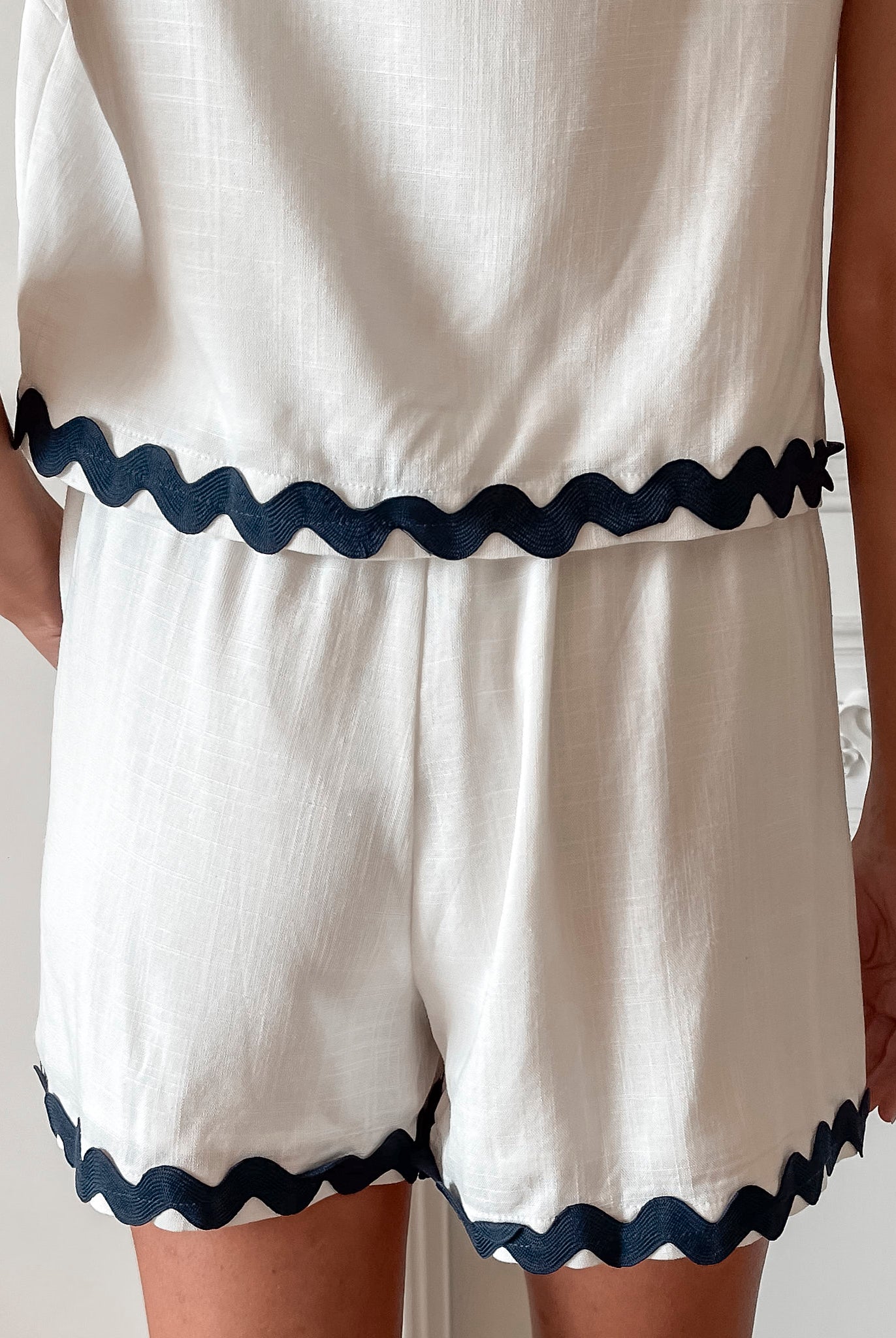 A woman is standing, wearing white high-waisted shorts with a string detail and side pockets, paired with a sleeveless top.