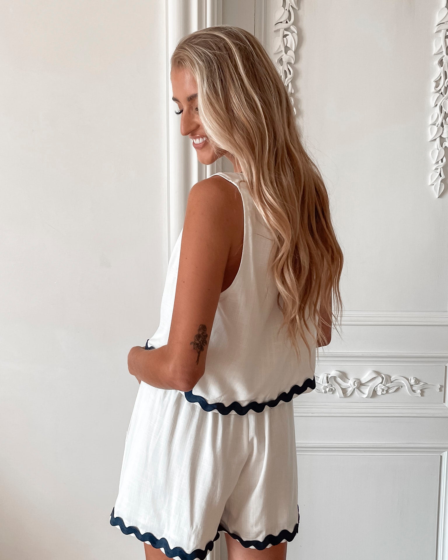 A woman is standing, wearing white high-waisted shorts with a string detail and side pockets, paired with a sleeveless top.