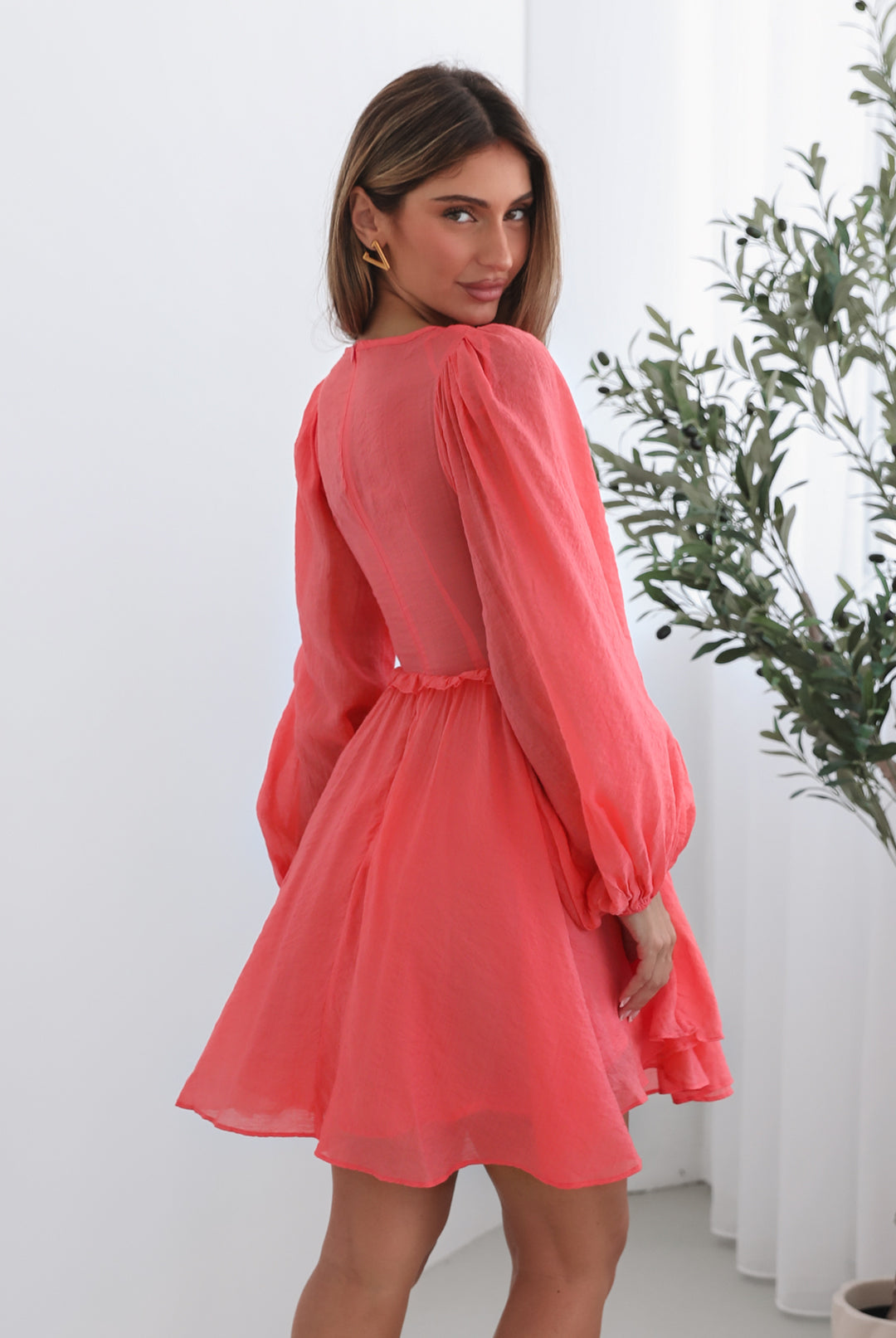 A woman wearing a coral-colored mini dress with a v-neckline, long bishop sleeves, smocked bodice, frills on waist, cut out, and an a-line skirt.