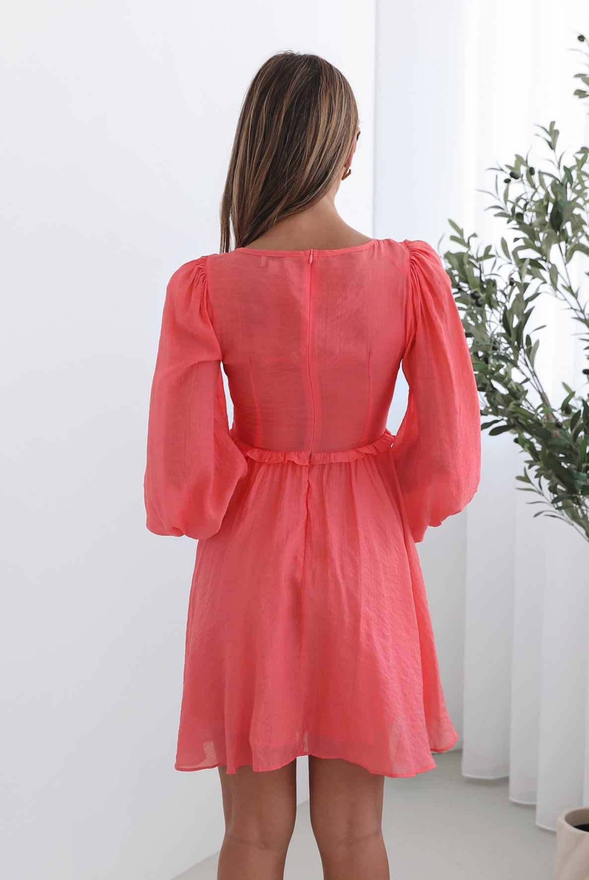 A woman wearing a coral-colored mini dress with a v-neckline, long bishop sleeves, smocked bodice, frills on waist, cut out, and an a-line skirt.