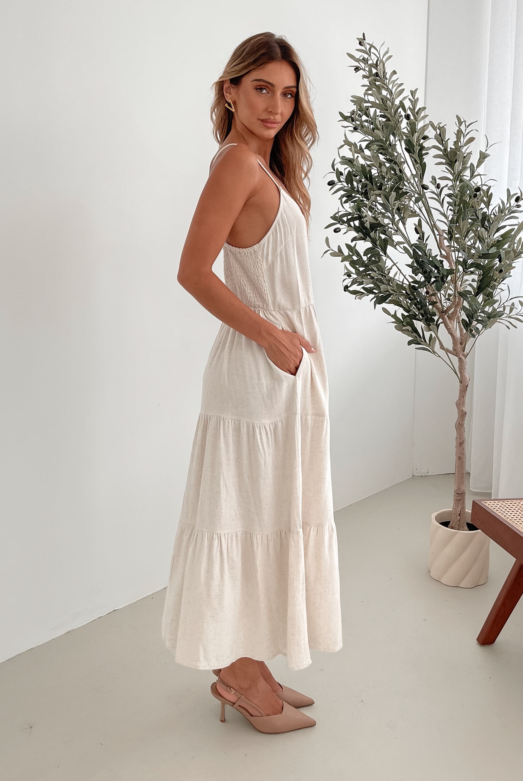 A woman wearing a beige, midi-length Sonya dress with a V neckline, adjustable spaghetti straps, a shirred back, and a tiered skirt. The dress is sleeveless and the photo shows the dress in a relaxed fit style.