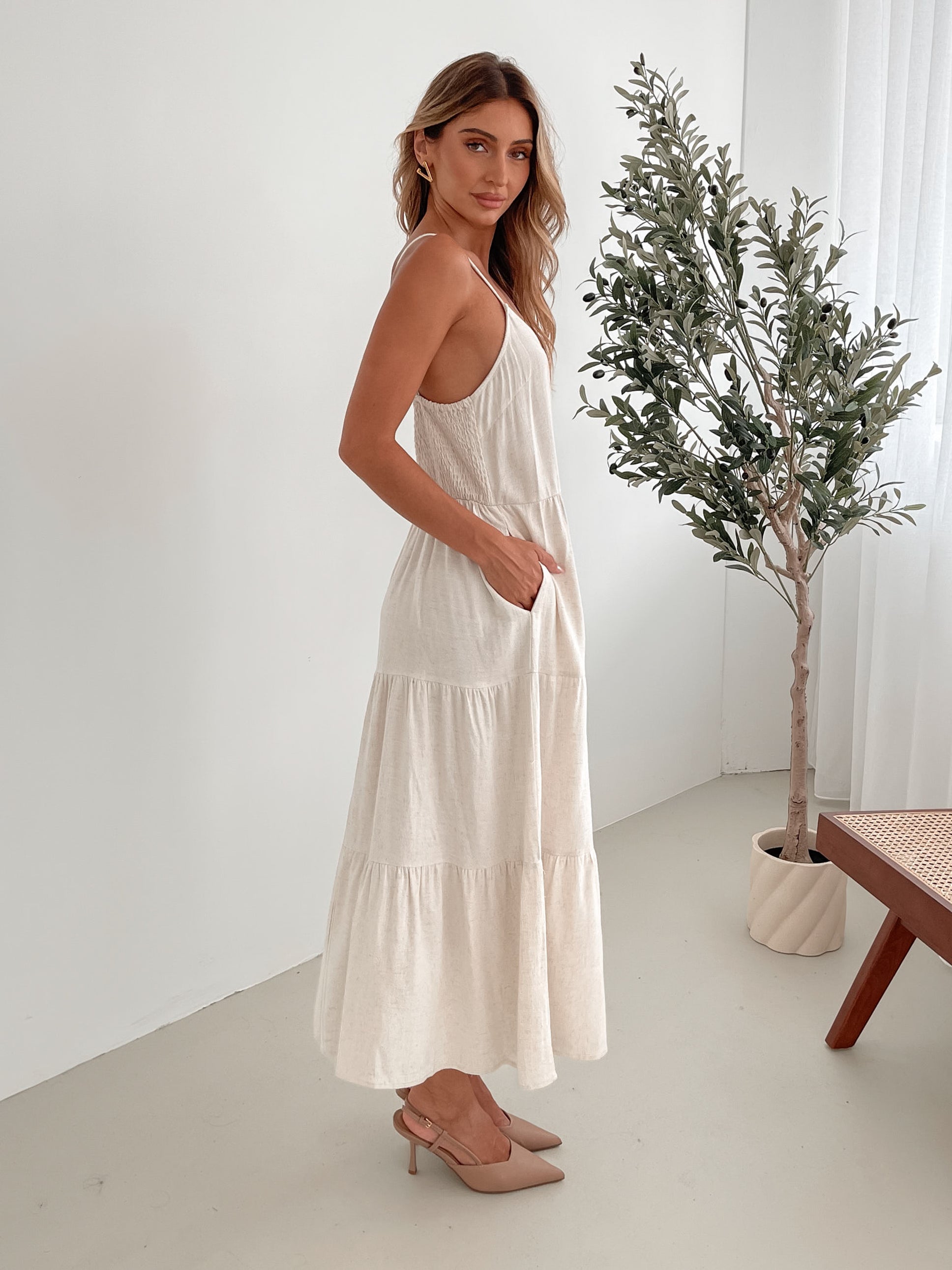 A woman wearing a beige, midi-length Sonya dress with a V neckline, adjustable spaghetti straps, a shirred back, and a tiered skirt. The dress is sleeveless and the photo shows the dress in a relaxed fit style.