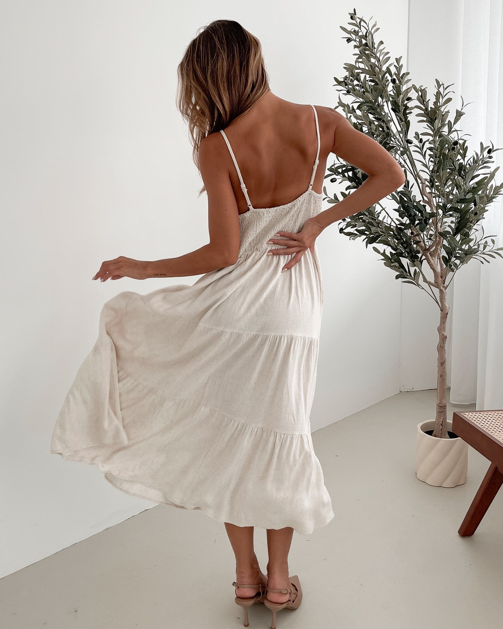 A woman wearing a beige, midi-length Sonya dress with a V neckline, adjustable spaghetti straps, a shirred back, and a tiered skirt. The dress is sleeveless and the photo shows the dress in a relaxed fit style.