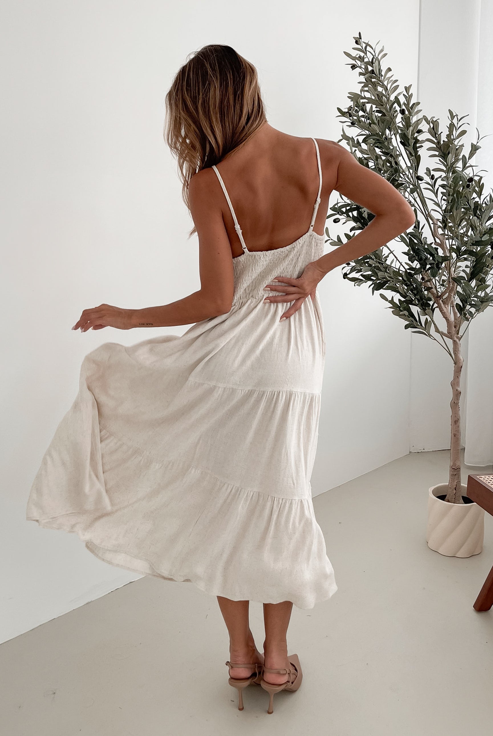 A woman wearing a beige, midi-length Sonya dress with a V neckline, adjustable spaghetti straps, a shirred back, and a tiered skirt. The dress is sleeveless and the photo shows the dress in a relaxed fit style.