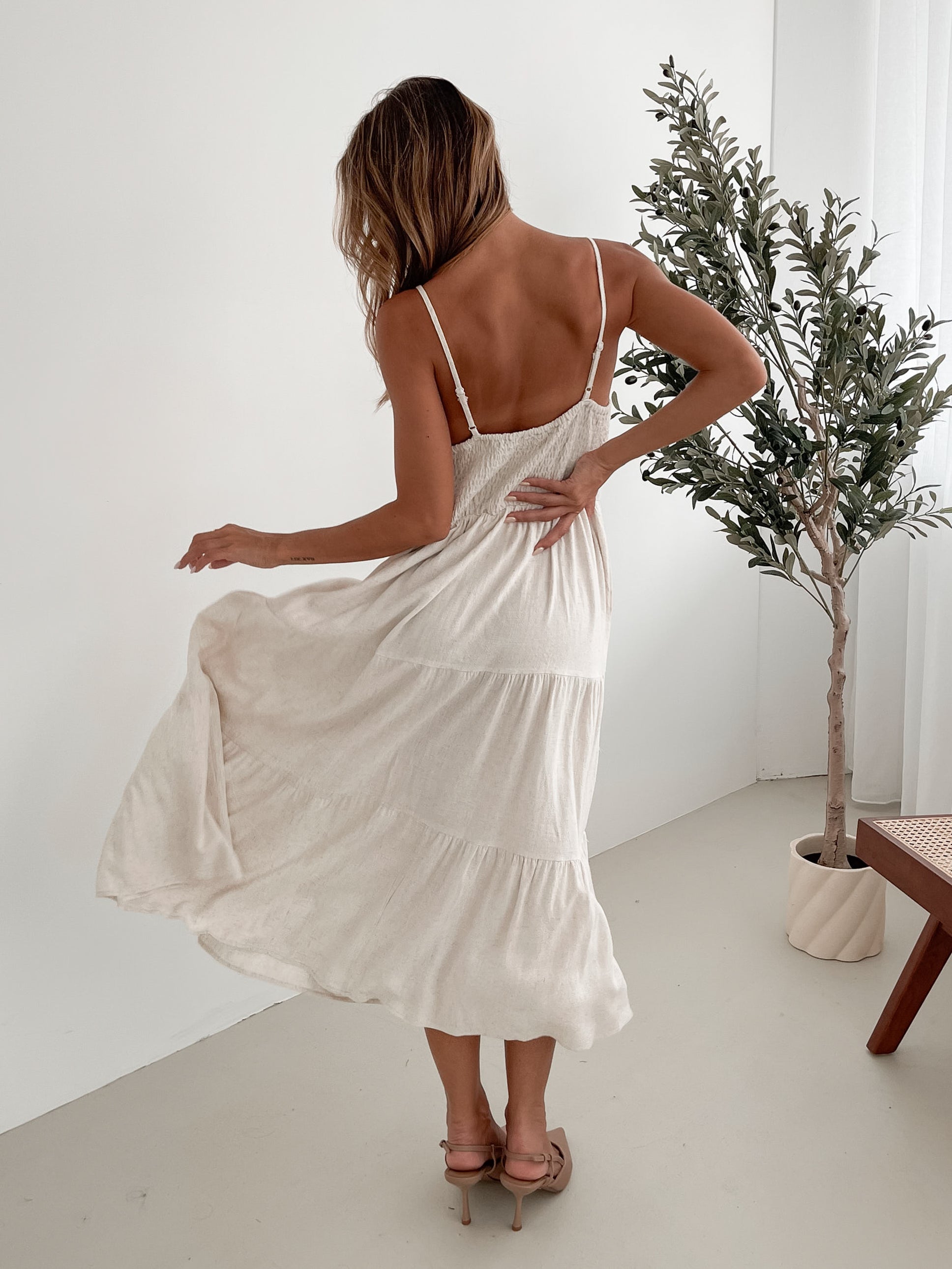 A woman wearing a beige, midi-length Sonya dress with a V neckline, adjustable spaghetti straps, a shirred back, and a tiered skirt. The dress is sleeveless and the photo shows the dress in a relaxed fit style.