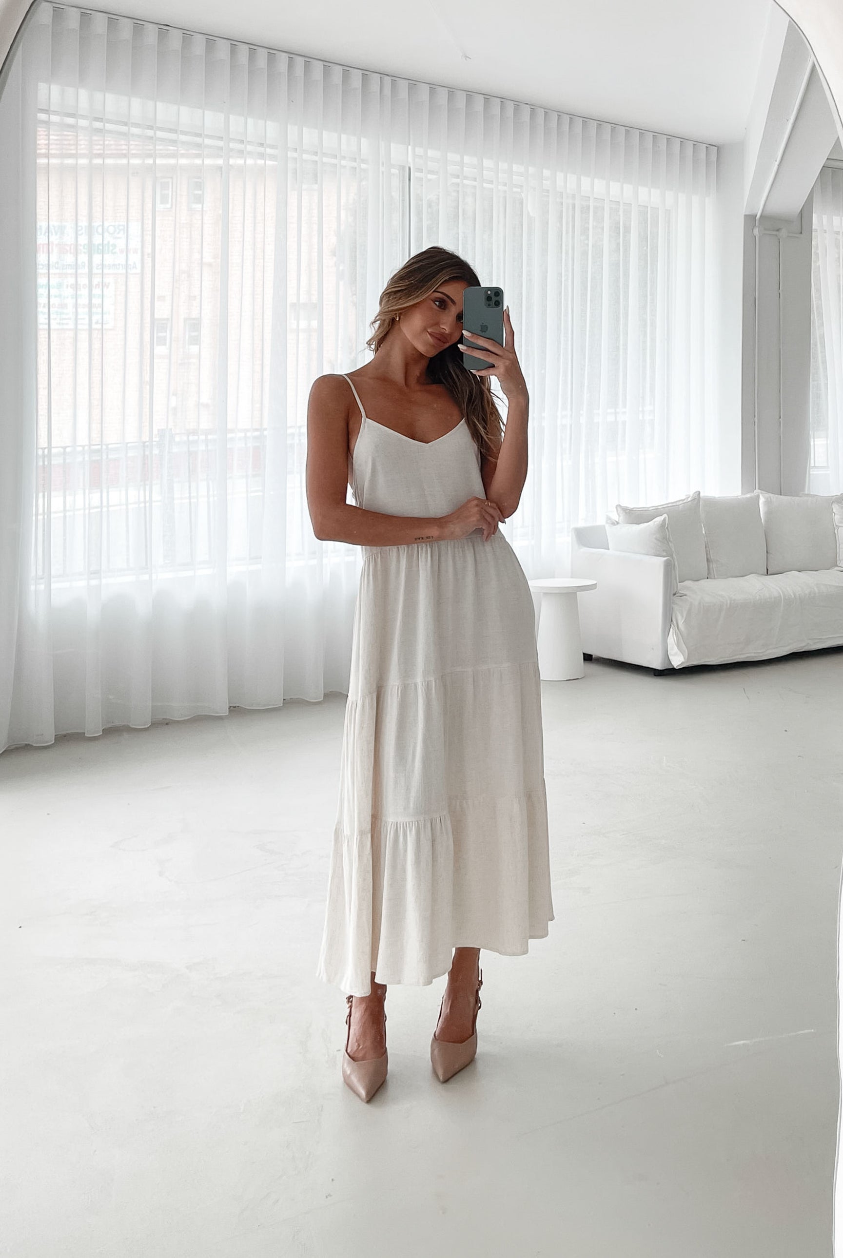 A woman wearing a beige, midi-length Sonya dress with a V neckline, adjustable spaghetti straps, a shirred back, and a tiered skirt. The dress is sleeveless and the photo shows the dress in a relaxed fit style.