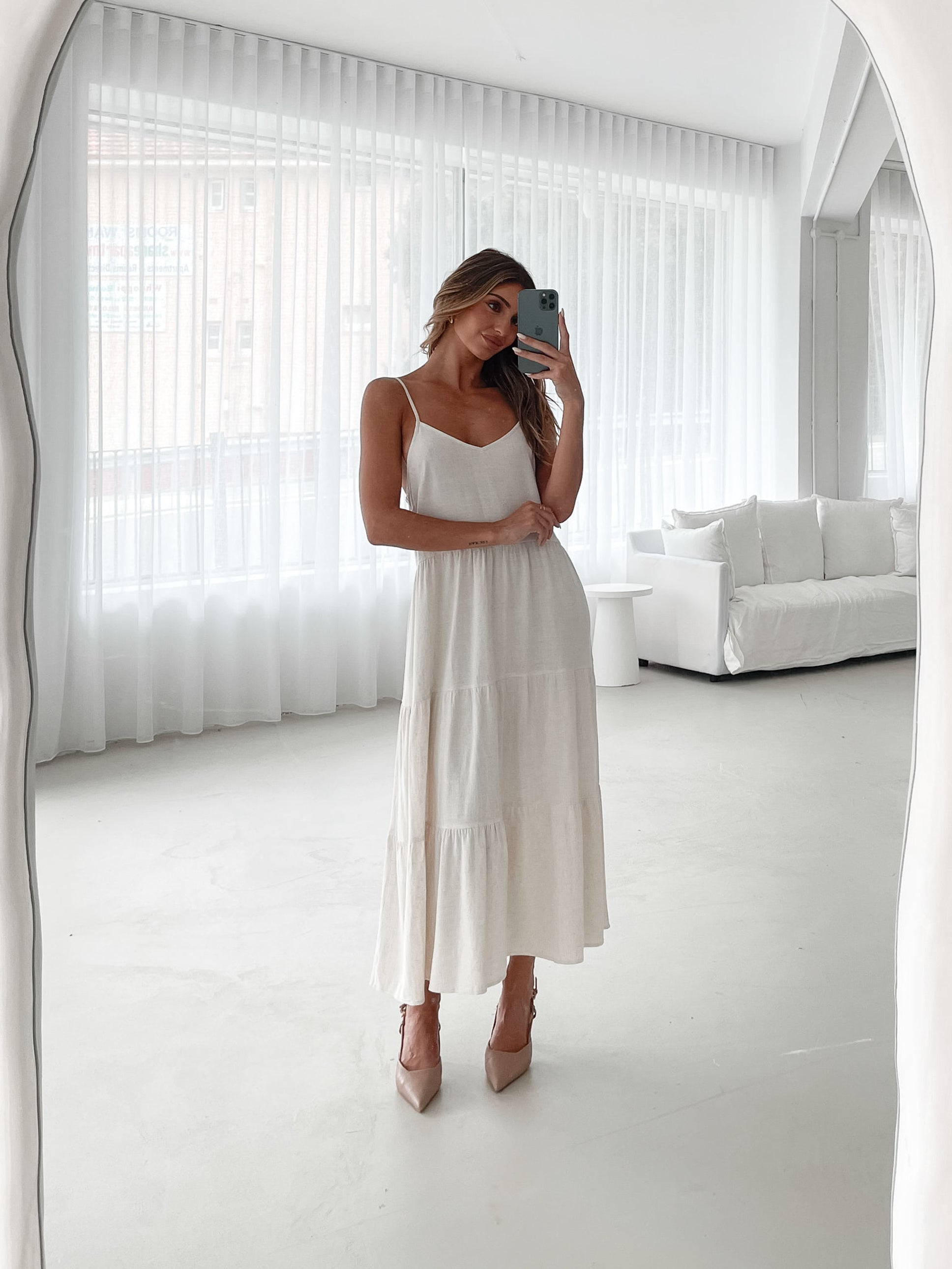 A woman wearing a beige, midi-length Sonya dress with a V neckline, adjustable spaghetti straps, a shirred back, and a tiered skirt. The dress is sleeveless and the photo shows the dress in a relaxed fit style.