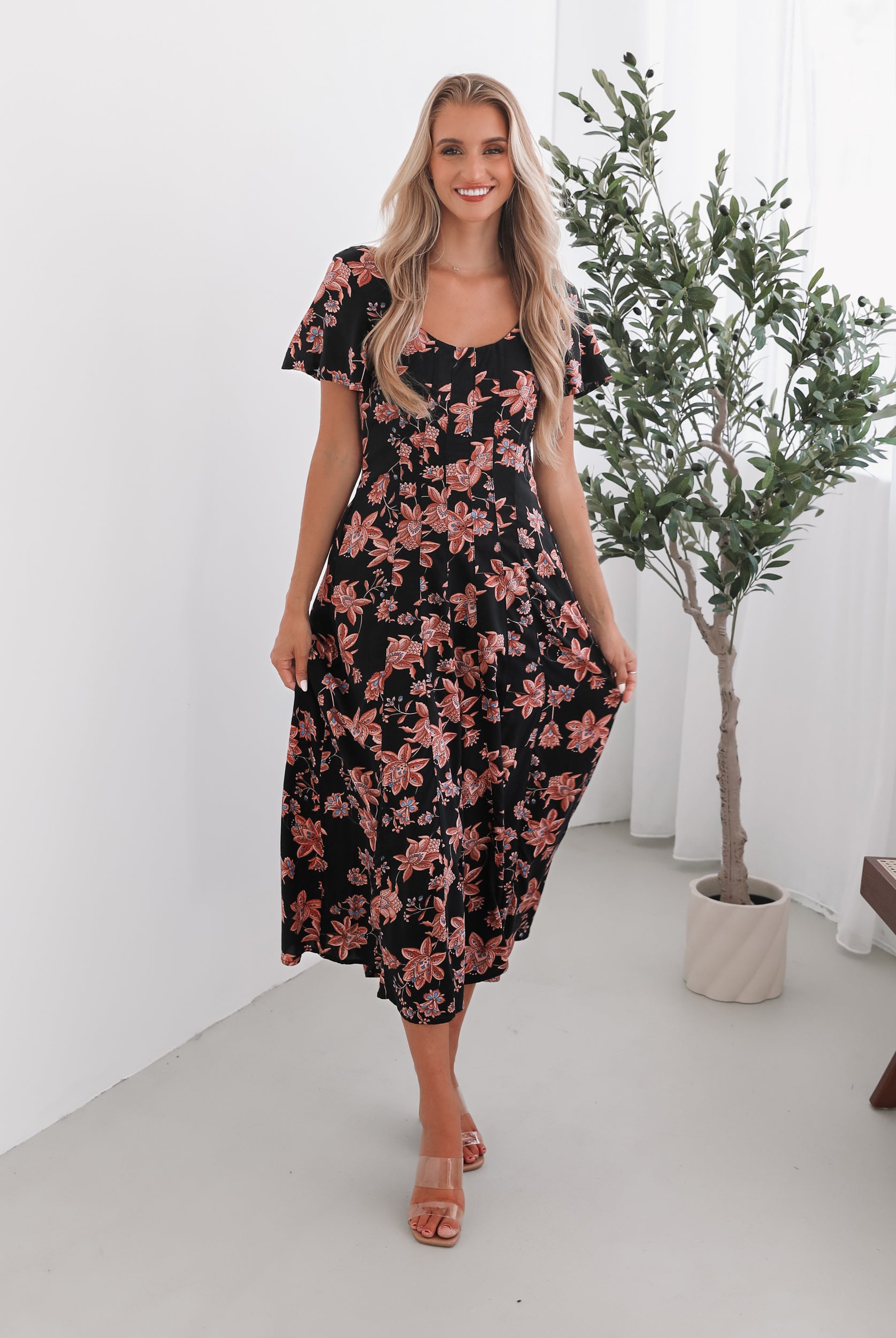 A woman standing in a room with a plant backdrop, wearing a black floral dress with a scoop neckline, short sleeves, and an a-line skirt.