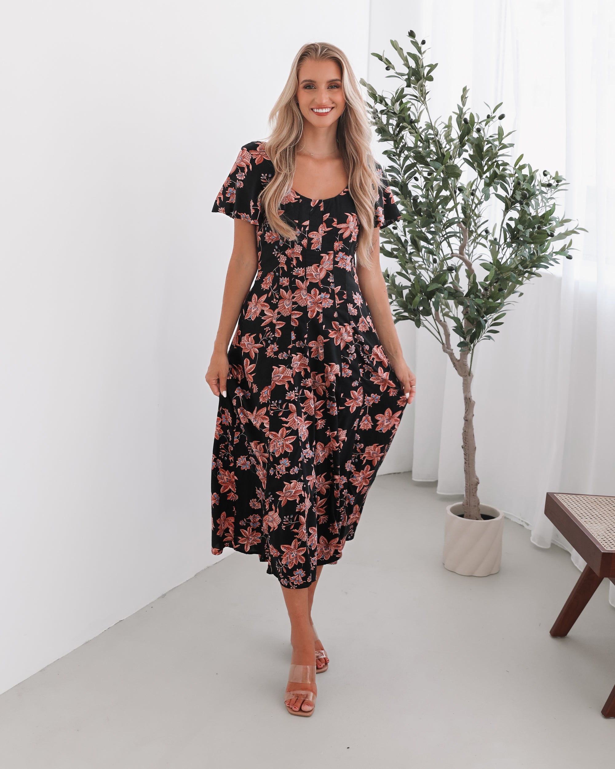 A woman standing in a room with a plant backdrop, wearing a black floral dress with a scoop neckline, short sleeves, and an a-line skirt.