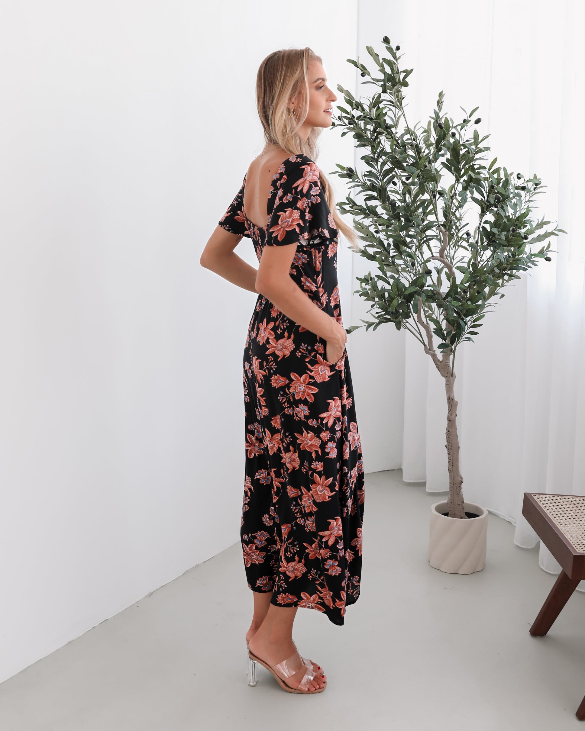 A woman standing in a room with a plant backdrop, wearing a black floral dress with a scoop neckline, short sleeves, and an a-line skirt.