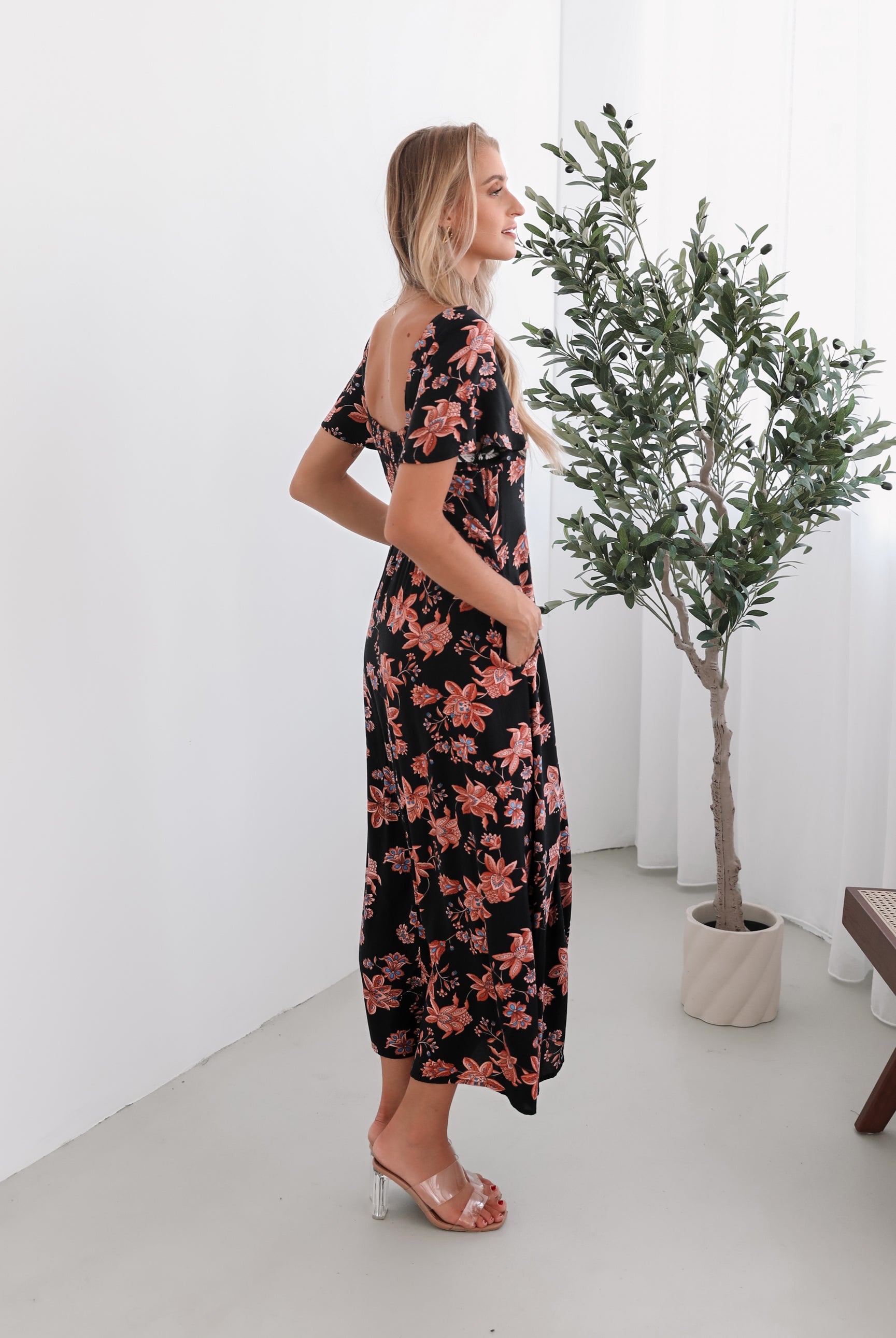 A woman standing in a room with a plant backdrop, wearing a black floral dress with a scoop neckline, short sleeves, and an a-line skirt.