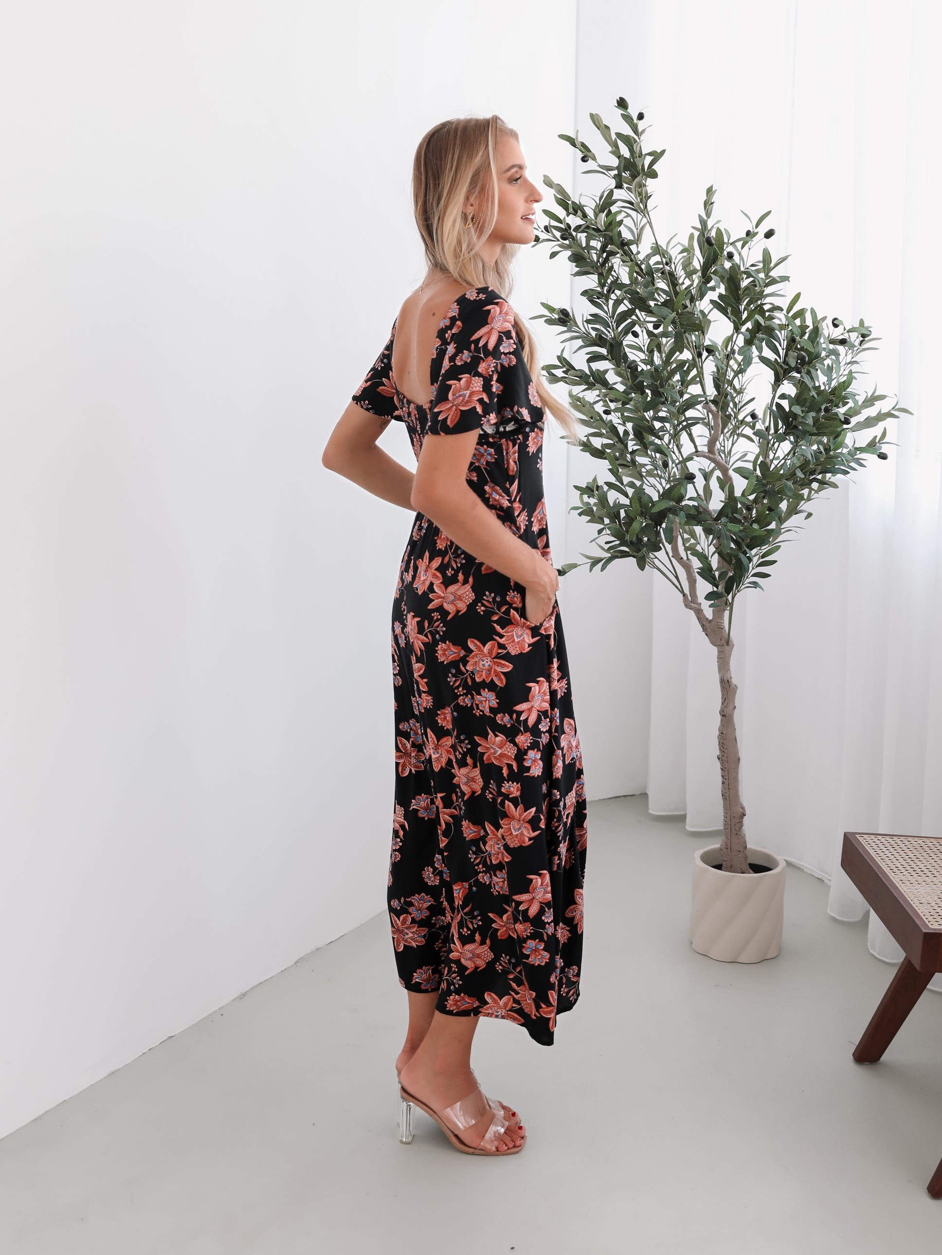 A woman standing in a room with a plant backdrop, wearing a black floral dress with a scoop neckline, short sleeves, and an a-line skirt.