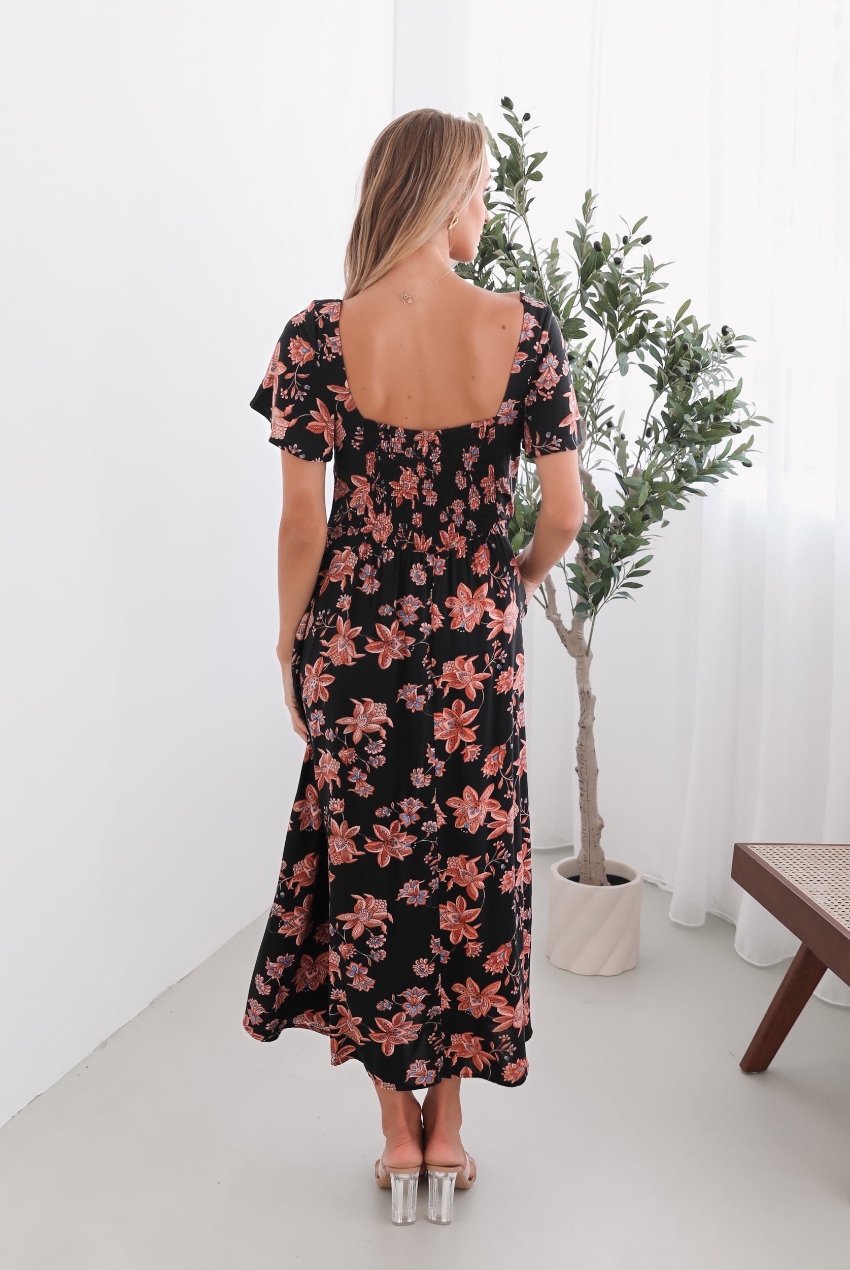 A woman standing in a room with a plant backdrop, wearing a black floral dress with a scoop neckline, short sleeves, and an a-line skirt.