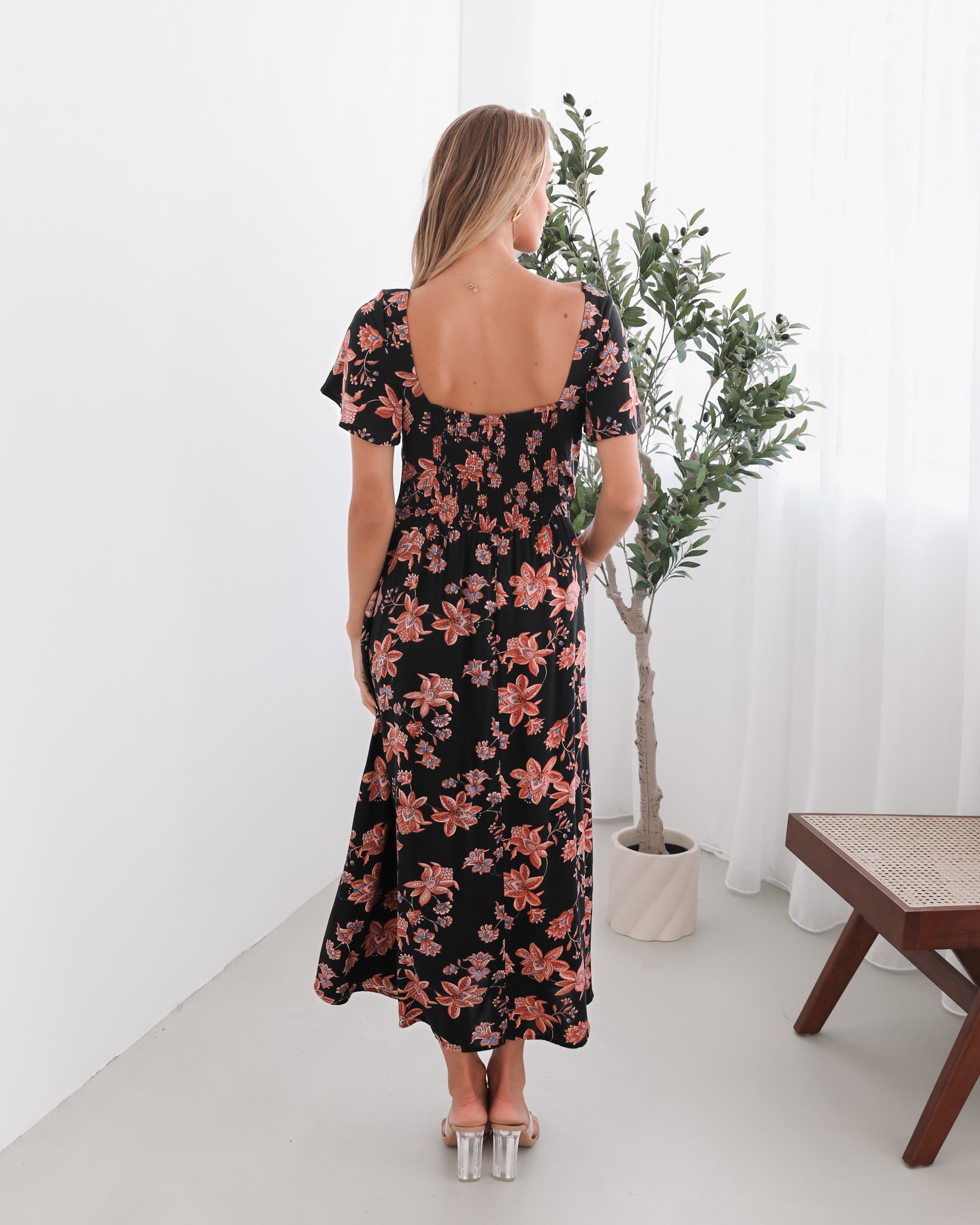 A woman standing in a room with a plant backdrop, wearing a black floral dress with a scoop neckline, short sleeves, and an a-line skirt.
