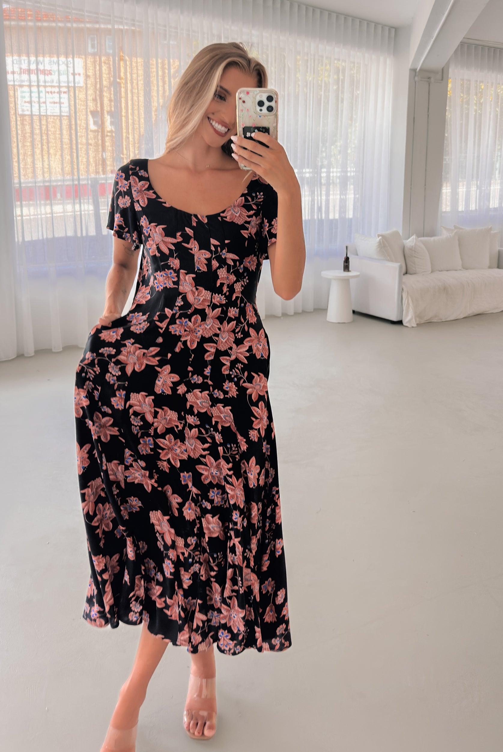 A woman standing in a room with a plant backdrop, wearing a black floral dress with a scoop neckline, short sleeves, and an a-line skirt.