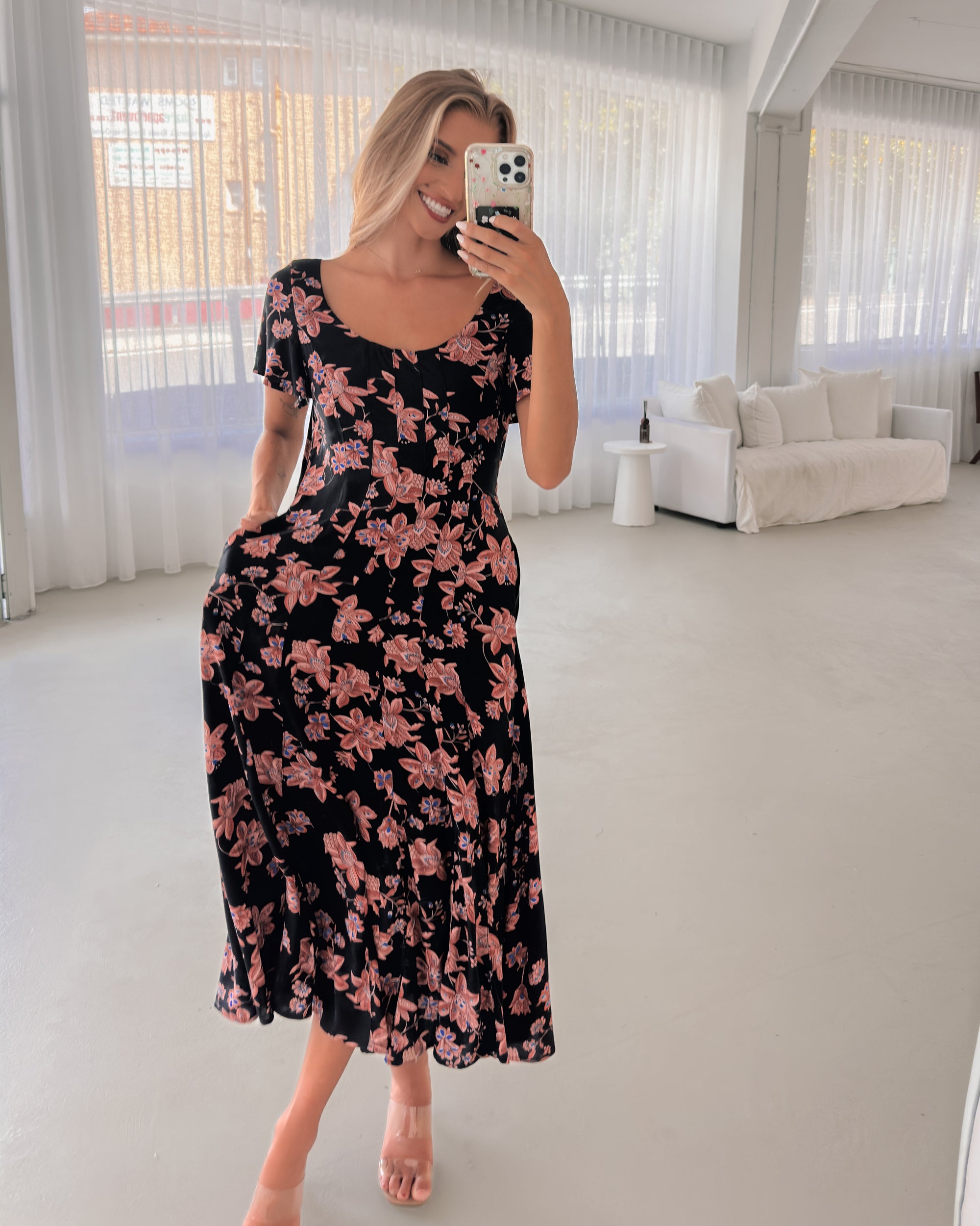 A woman standing in a room with a plant backdrop, wearing a black floral dress with a scoop neckline, short sleeves, and an a-line skirt.