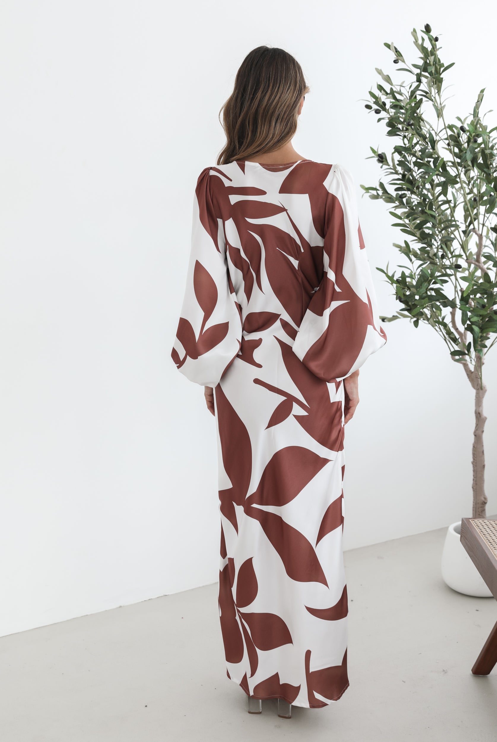 A model wearing a white and camel colored maxi dress with a wide v-neckline and long bishop sleeves, in a fit-to-flare silhouette with a maxi length hem.