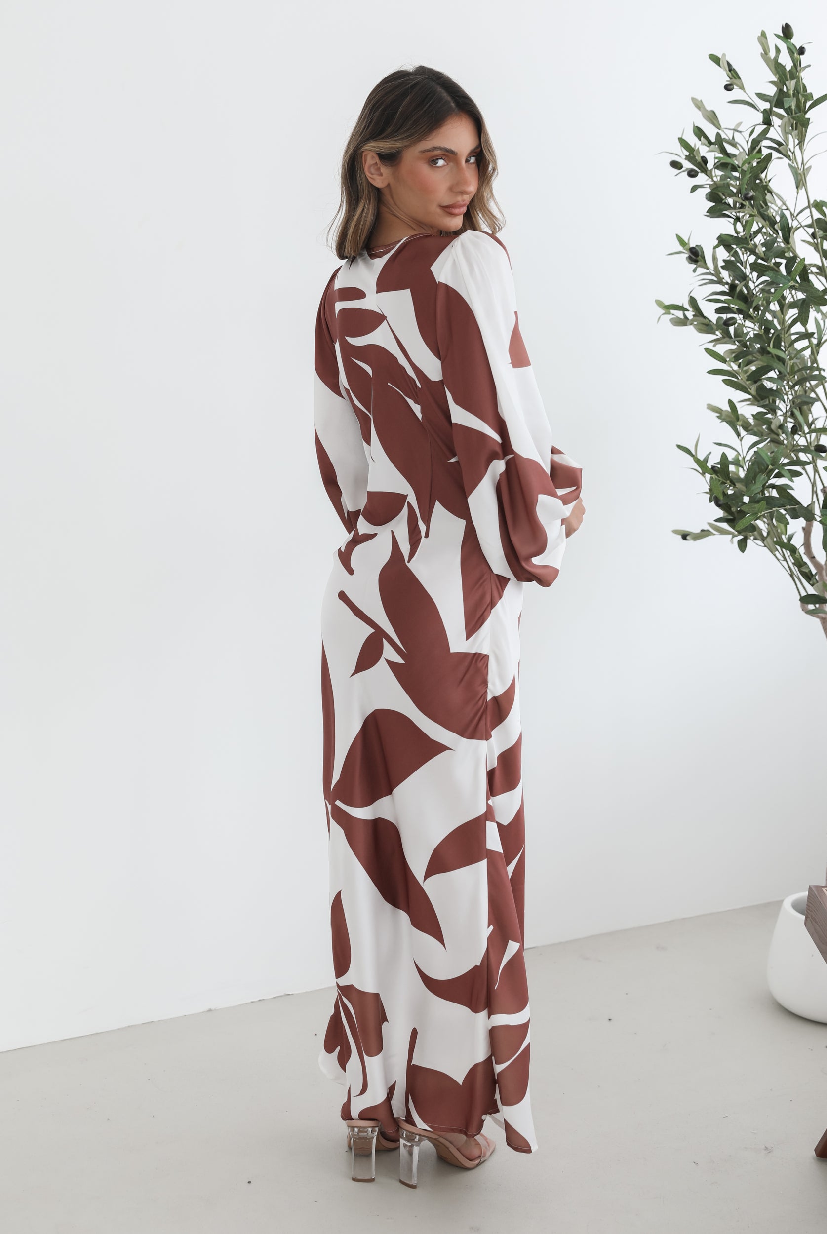 A model wearing a white and camel colored maxi dress with a wide v-neckline and long bishop sleeves, in a fit-to-flare silhouette with a maxi length hem.