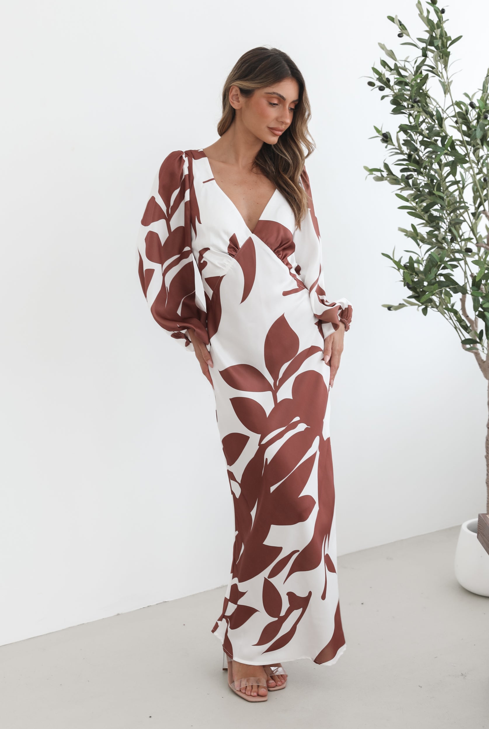 A model wearing a white and camel colored maxi dress with a wide v-neckline and long bishop sleeves, in a fit-to-flare silhouette with a maxi length hem.