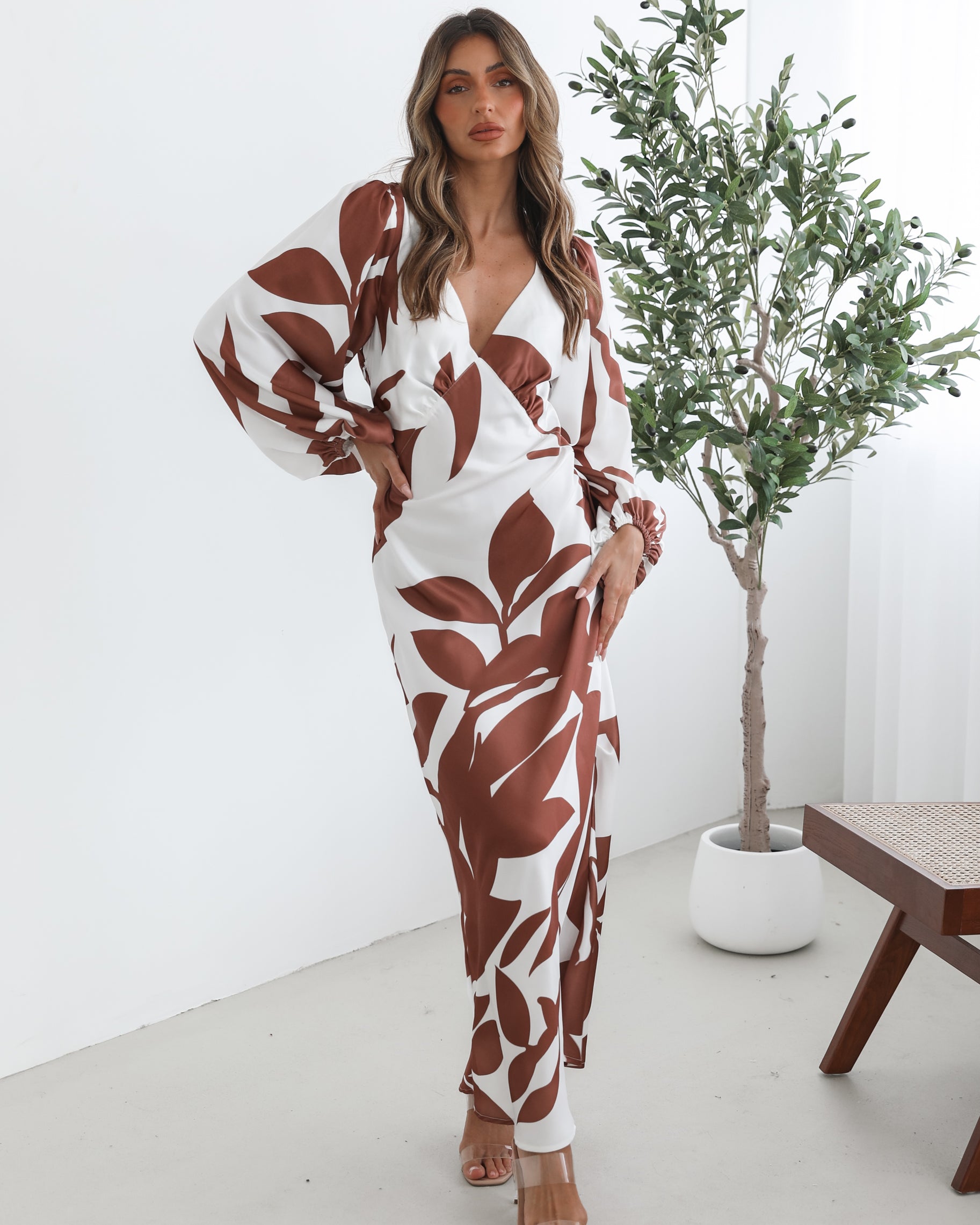 A model wearing a white and camel colored maxi dress with a wide v-neckline and long bishop sleeves, in a fit-to-flare silhouette with a maxi length hem.