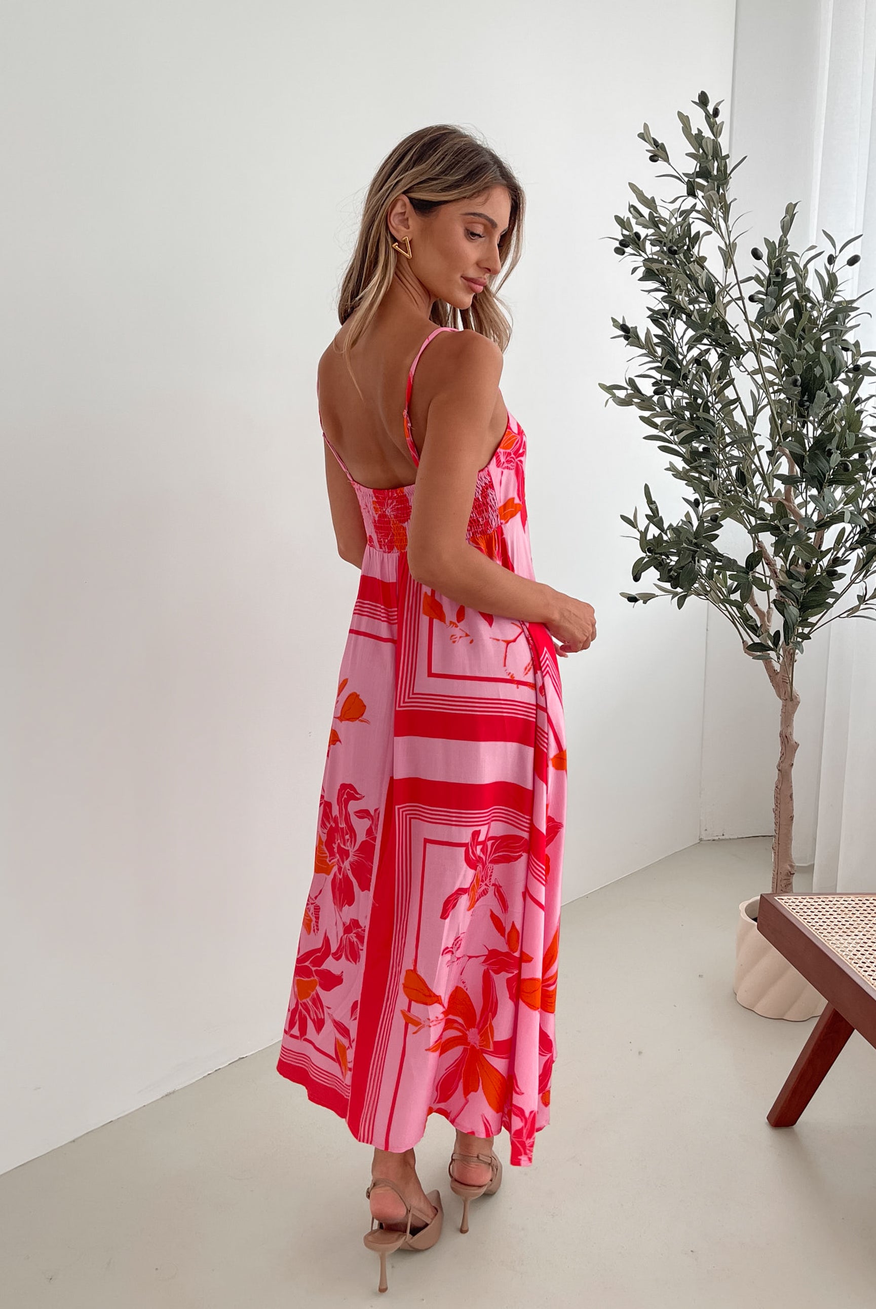 A woman wearing a pink floral dress with spaghetti straps and a square neckline. The dress has a flowy a-line skirt and she is standing in a room with a plant in the background.