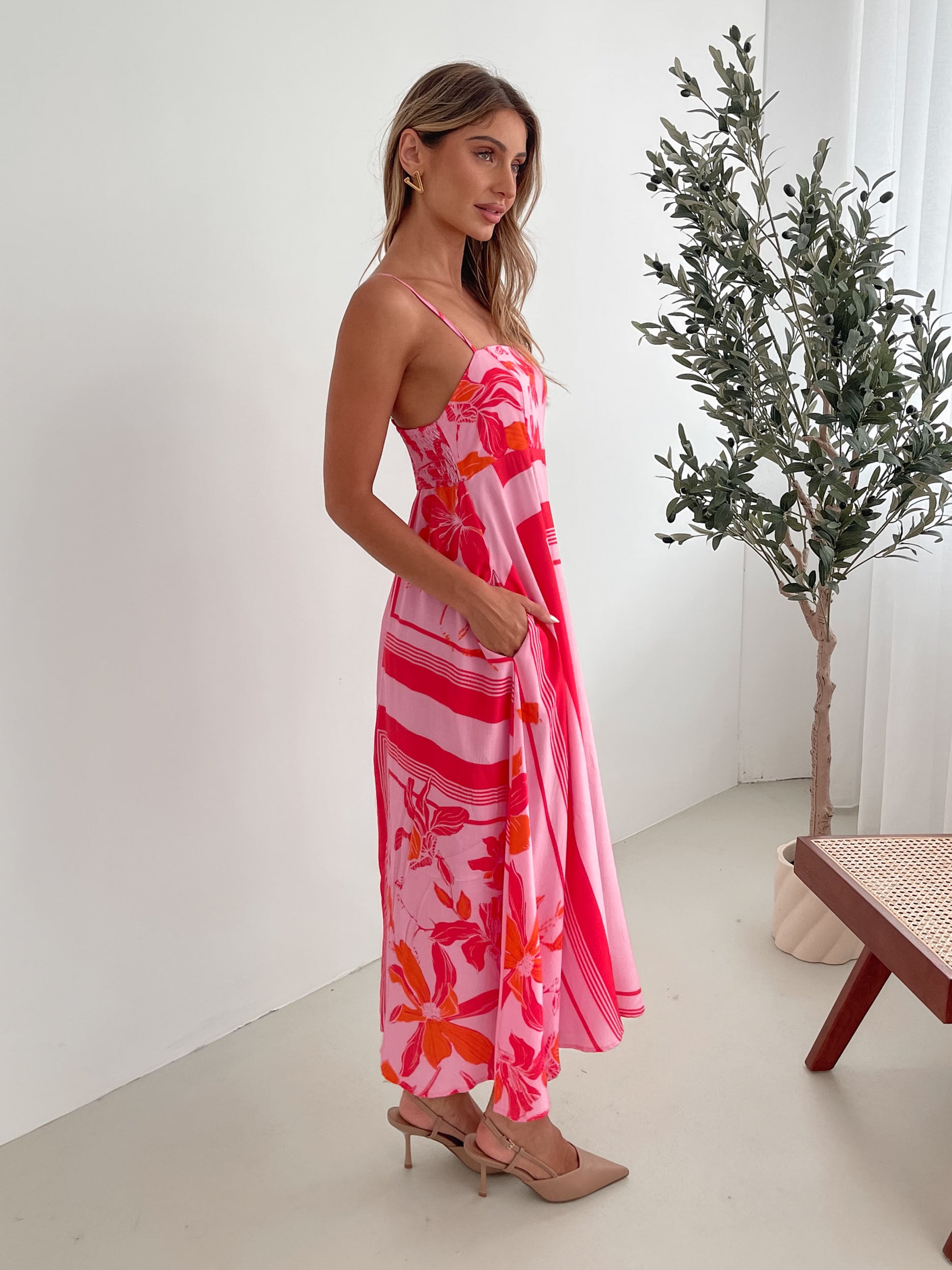 A woman wearing a pink floral dress with spaghetti straps and a square neckline. The dress has a flowy a-line skirt and she is standing in a room with a plant in the background.