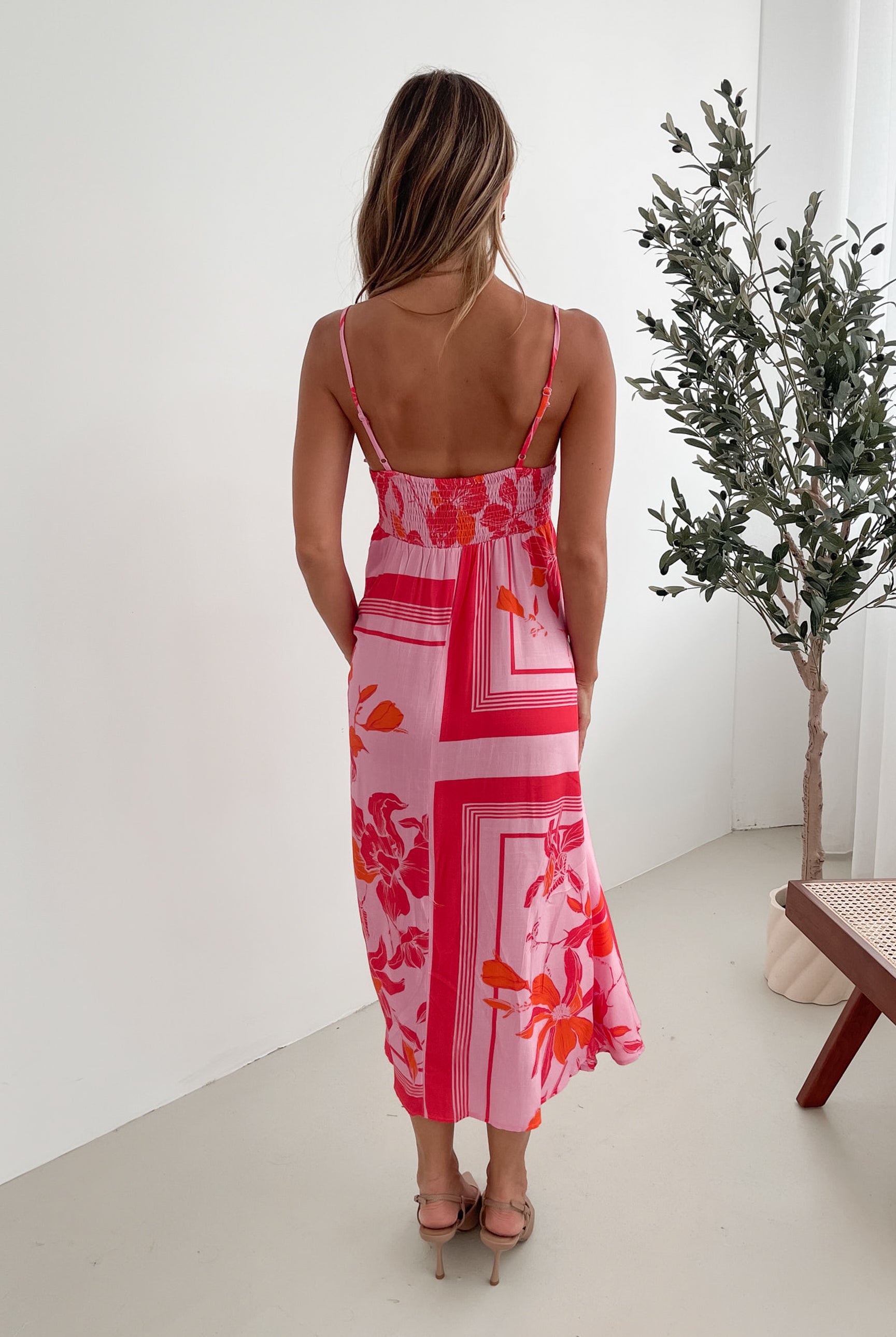 A woman wearing a pink floral dress with spaghetti straps and a square neckline. The dress has a flowy a-line skirt and she is standing in a room with a plant in the background.