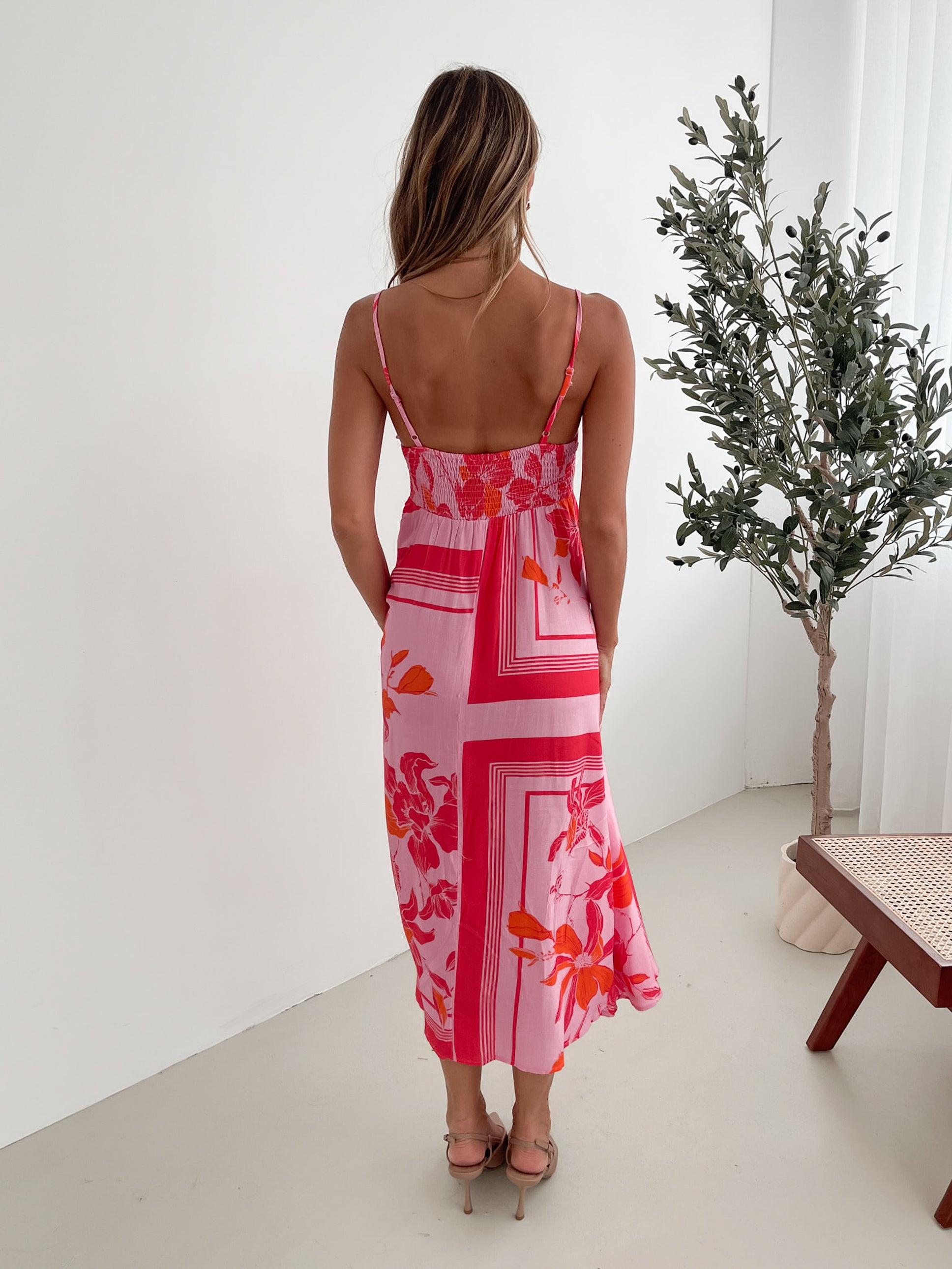 A woman wearing a pink floral dress with spaghetti straps and a square neckline. The dress has a flowy a-line skirt and she is standing in a room with a plant in the background.