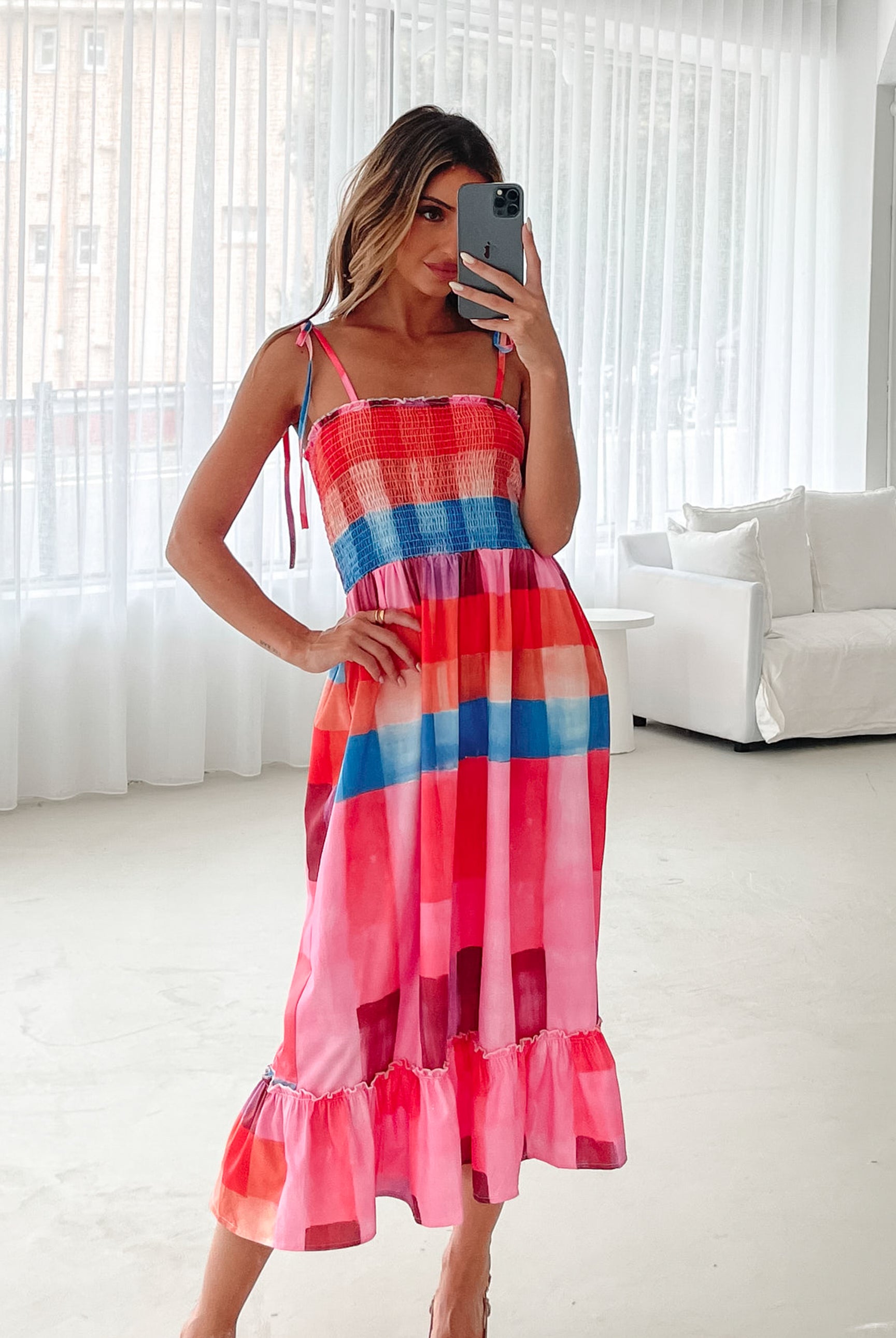 A woman standing in a room with a white wall and a small wooden chair, wearing a multicolored sleeveless dress with a square neckline and spaghetti straps. The dress features a smocked bodice and a flowy skirt.
