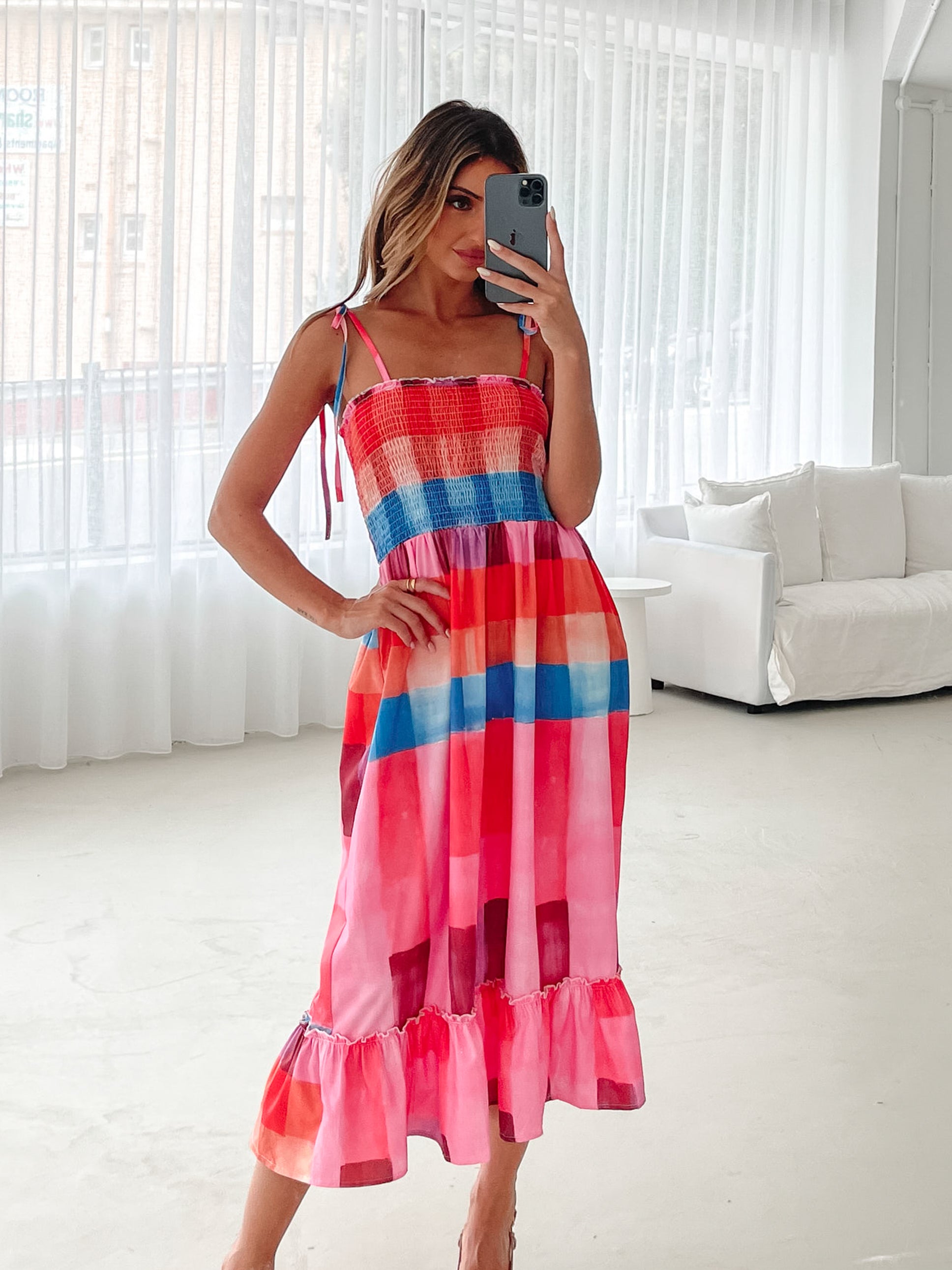 A woman standing in a room with a white wall and a small wooden chair, wearing a multicolored sleeveless dress with a square neckline and spaghetti straps. The dress features a smocked bodice and a flowy skirt.