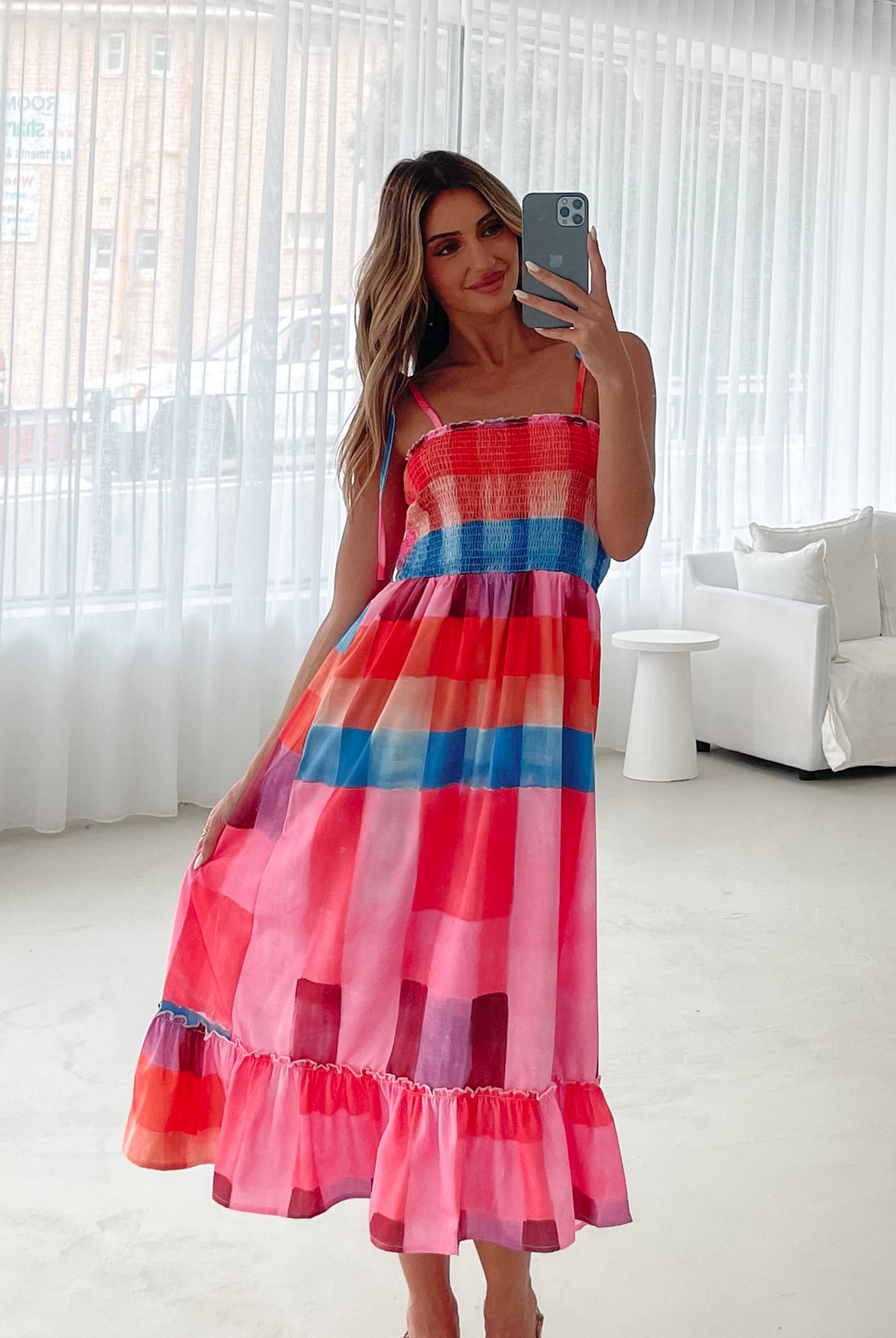 A woman standing in a room with a white wall and a small wooden chair, wearing a multicolored sleeveless dress with a square neckline and spaghetti straps. The dress features a smocked bodice and a flowy skirt.