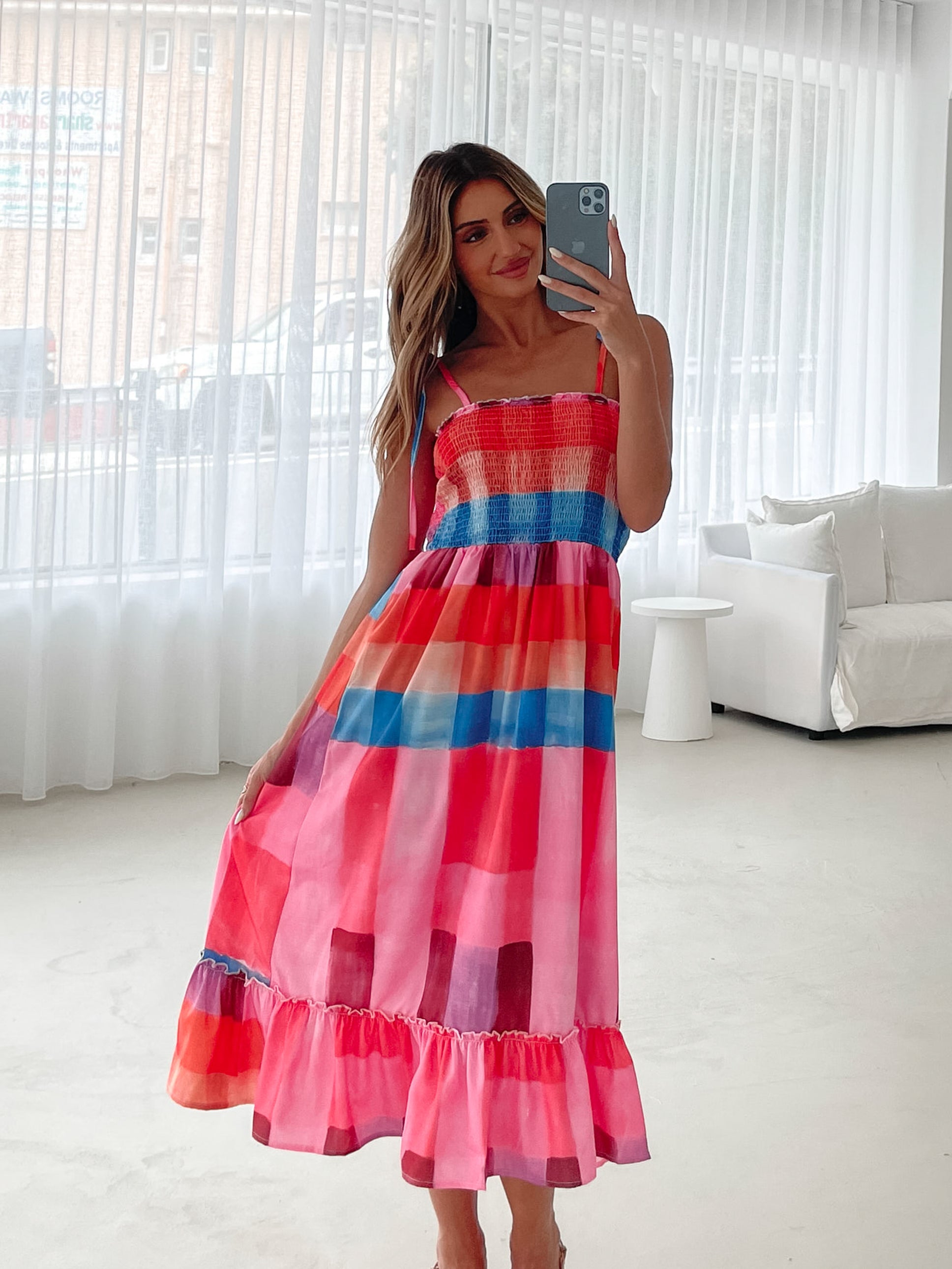 A woman standing in a room with a white wall and a small wooden chair, wearing a multicolored sleeveless dress with a square neckline and spaghetti straps. The dress features a smocked bodice and a flowy skirt.