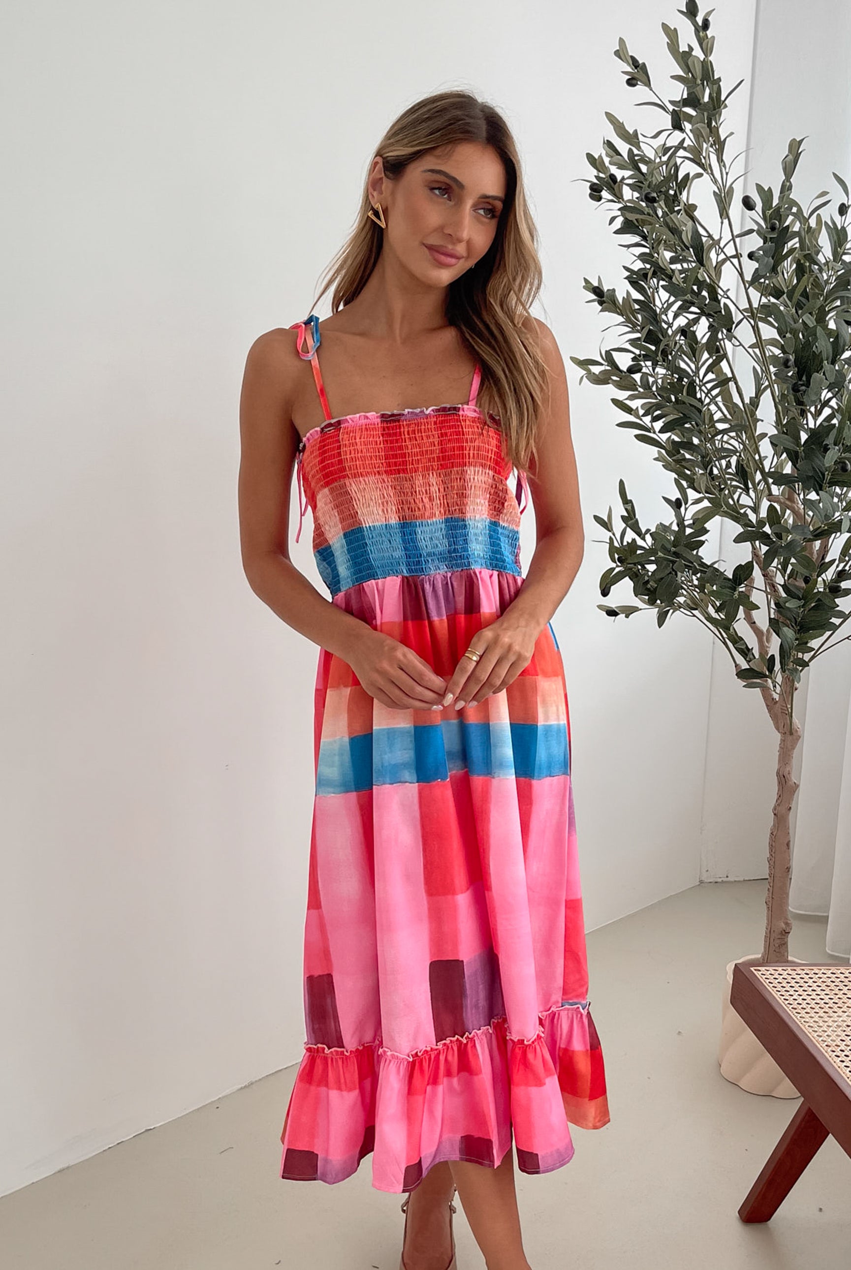 A woman standing in a room with a white wall and a small wooden chair, wearing a multicolored sleeveless dress with a square neckline and spaghetti straps. The dress features a smocked bodice and a flowy skirt.