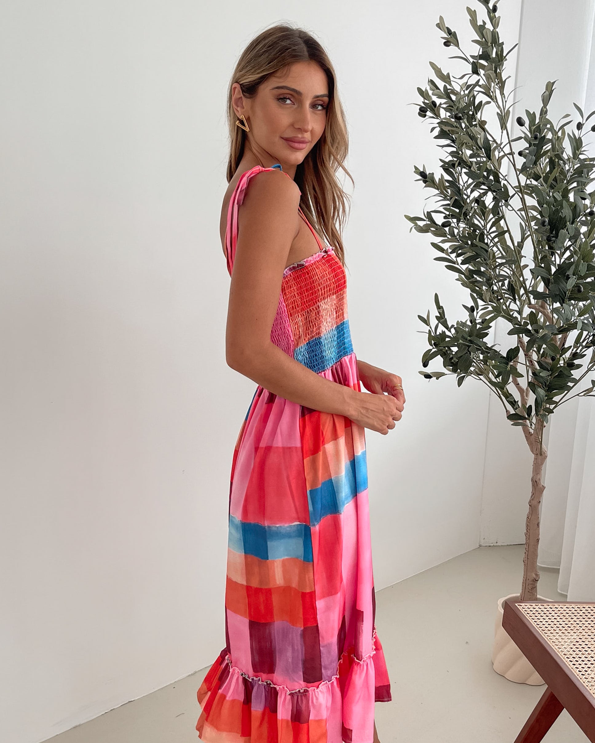 A woman standing in a room with a white wall and a small wooden chair, wearing a multicolored sleeveless dress with a square neckline and spaghetti straps. The dress features a smocked bodice and a flowy skirt.