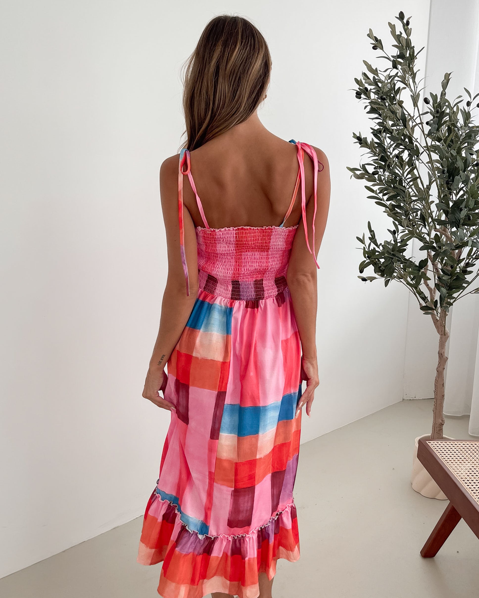 A woman standing in a room with a white wall and a small wooden chair, wearing a multicolored sleeveless dress with a square neckline and spaghetti straps. The dress features a smocked bodice and a flowy skirt.