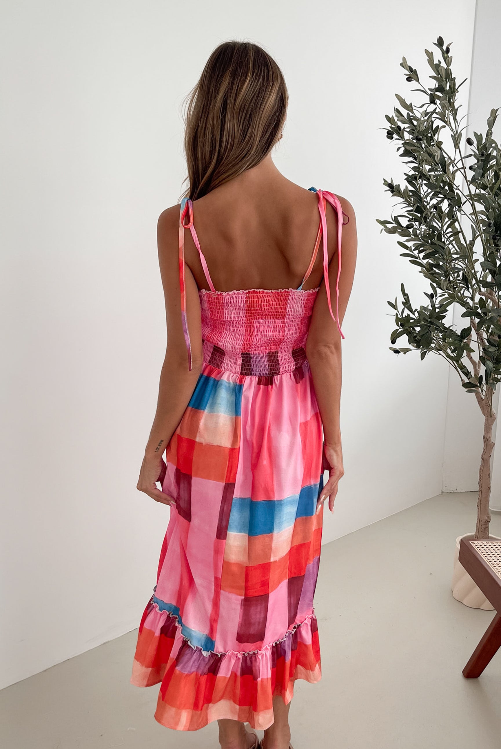 A woman standing in a room with a white wall and a small wooden chair, wearing a multicolored sleeveless dress with a square neckline and spaghetti straps. The dress features a smocked bodice and a flowy skirt.
