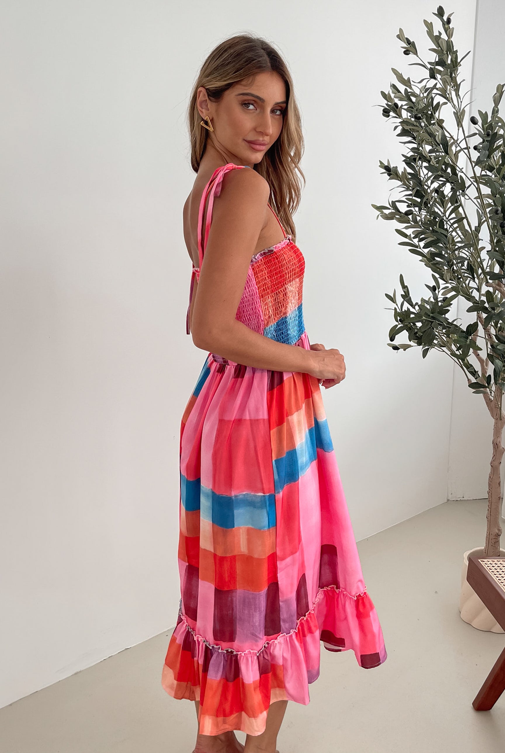 A woman standing in a room with a white wall and a small wooden chair, wearing a multicolored sleeveless dress with a square neckline and spaghetti straps. The dress features a smocked bodice and a flowy skirt.