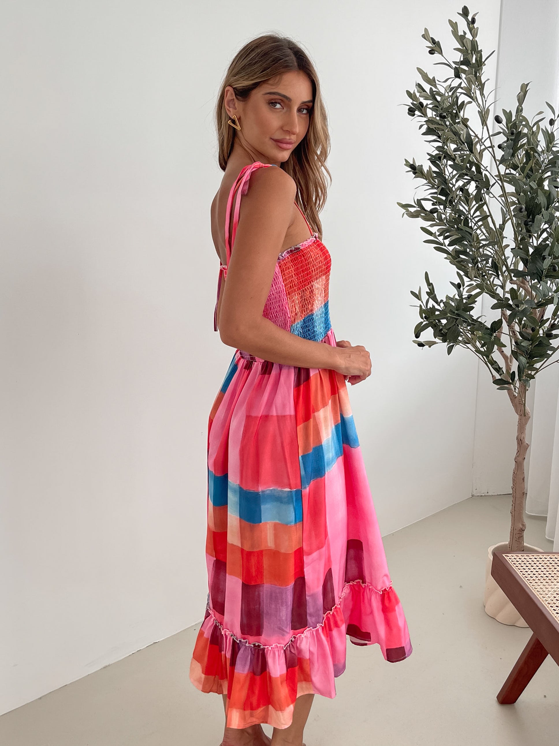 A woman standing in a room with a white wall and a small wooden chair, wearing a multicolored sleeveless dress with a square neckline and spaghetti straps. The dress features a smocked bodice and a flowy skirt.