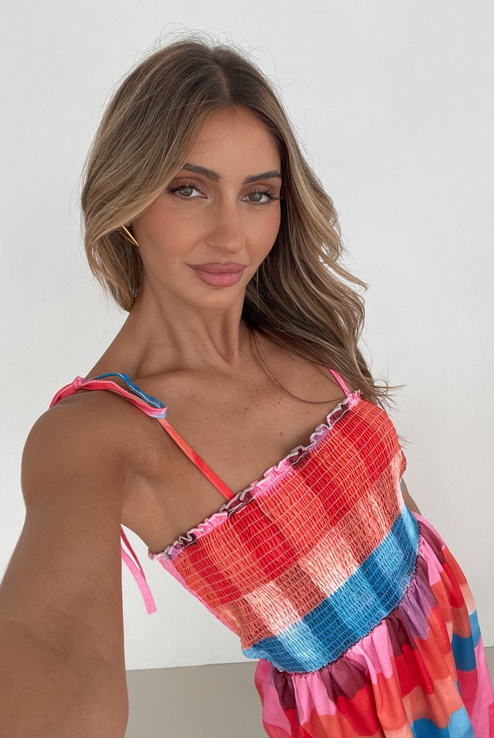 A woman standing in a room with a white wall and a small wooden chair, wearing a multicolored sleeveless dress with a square neckline and spaghetti straps. The dress features a smocked bodice and a flowy skirt.