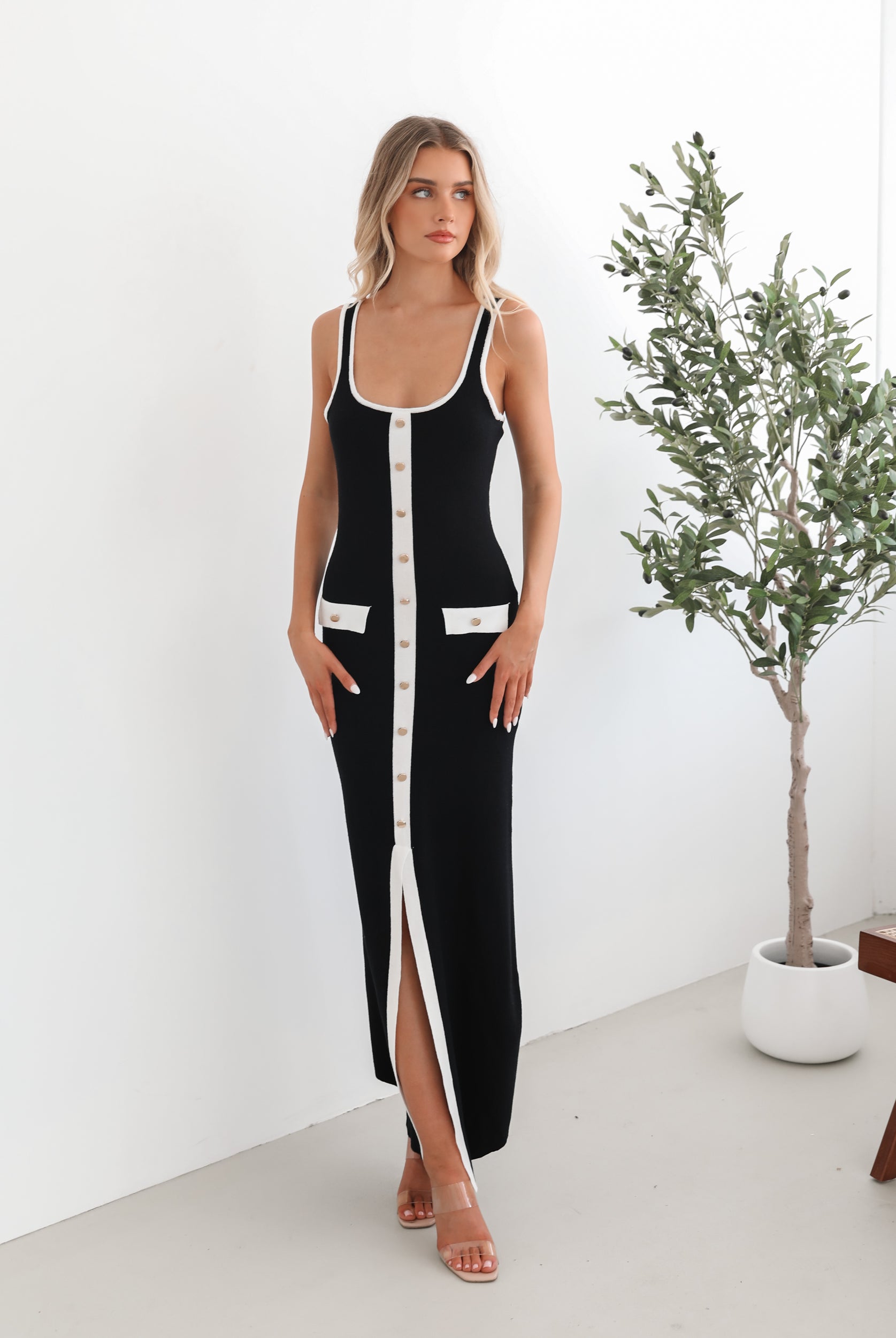 A model wearing a black sleeveless knit dress with a scoop neckline, center-front buttons, and a maxi length hem.