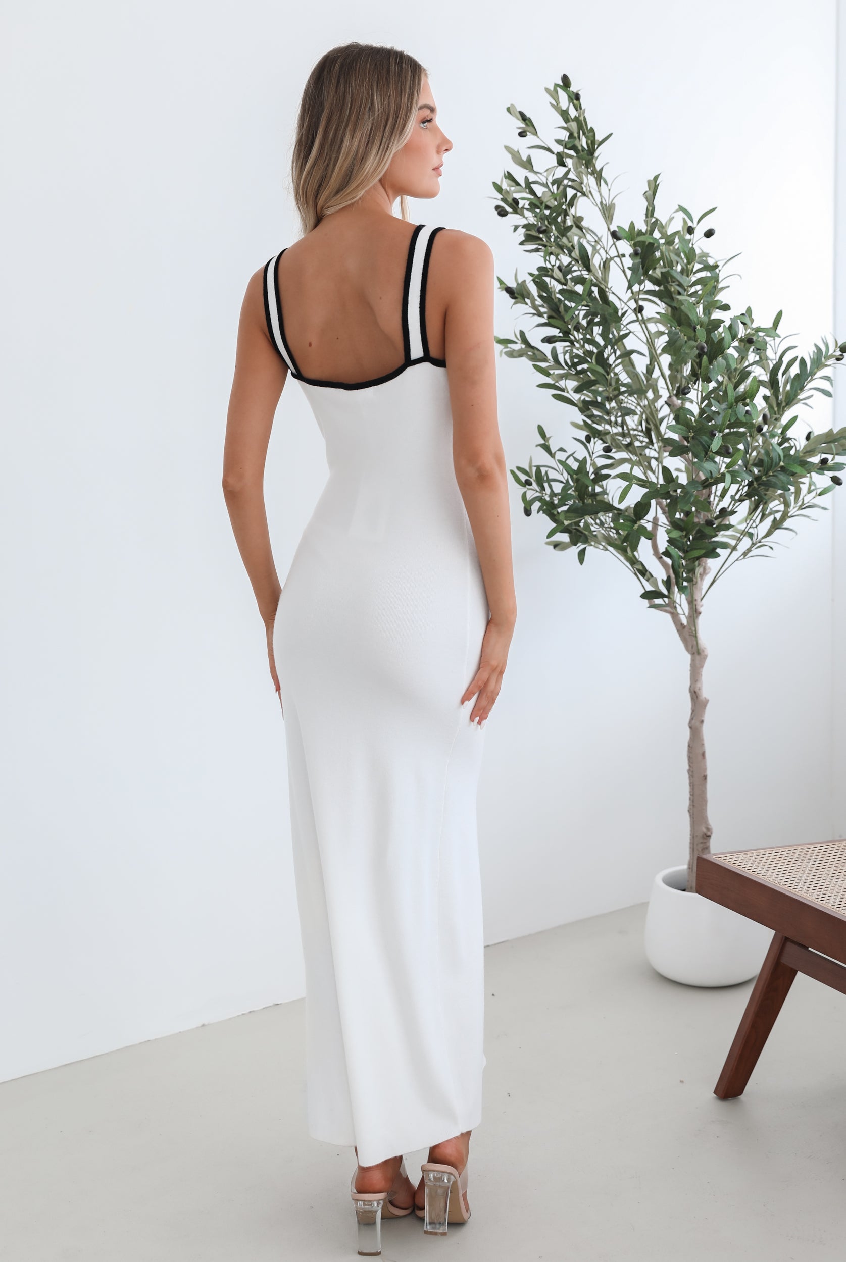 A woman wearing a white knit dress with a scoop neckline, center-front buttons, and a maxi length hem. The dress features a sleeveless design and a split at the front.