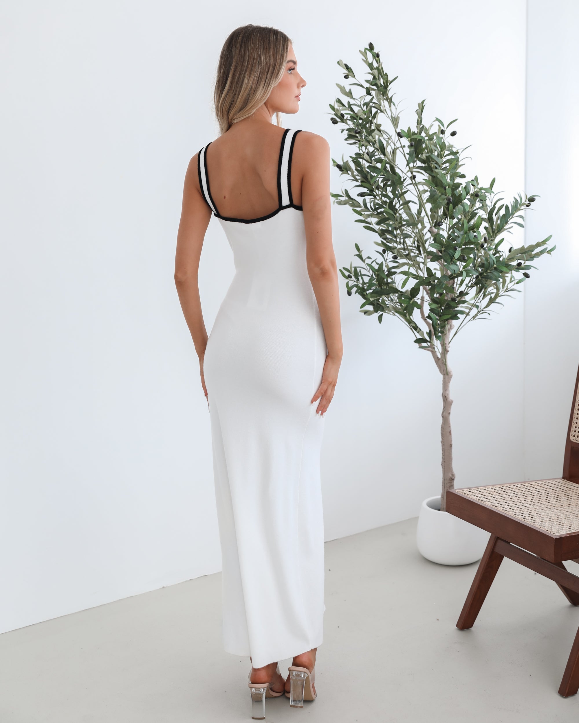 A woman wearing a white knit dress with a scoop neckline, center-front buttons, and a maxi length hem. The dress features a sleeveless design and a split at the front.