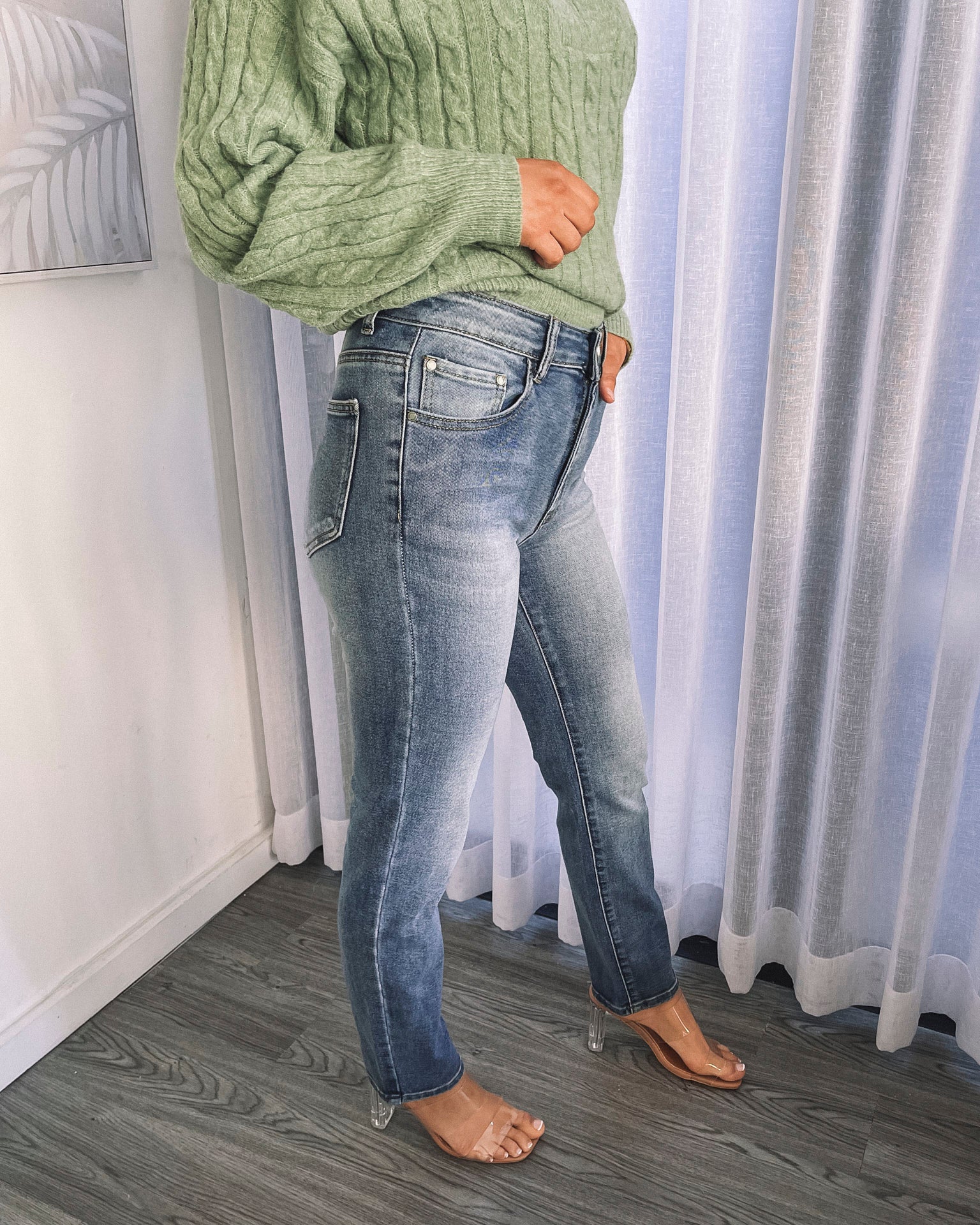 A person wearing mid-wash blue denim jeans and a green sweater, standing in a room with white curtains.